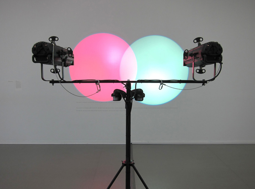 Two overhead lights project intersecting red and cyan circles onto a gray wall, mounted on a black tripod stand. The circles create a blend of light pink where they overlap. This is an AI generated description. To give feedback, email ai@moma.org.