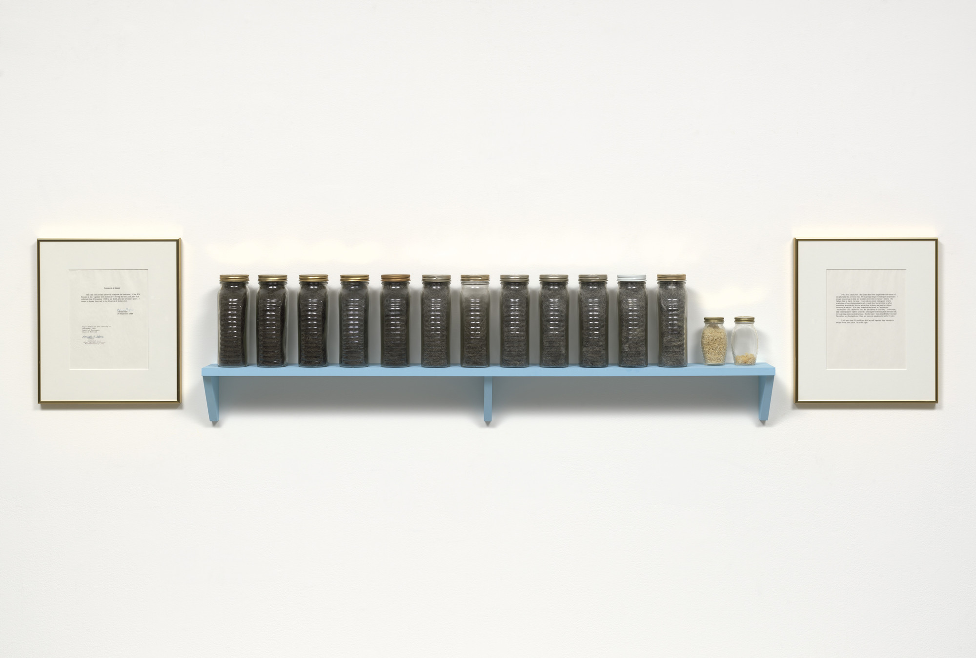 A light blue shelf holds 13 jars with varying contents, set against a plain white wall. Two framed papers, one on each side of the shelf, feature typed text. This is an AI generated description. To give feedback, email ai@moma.org.