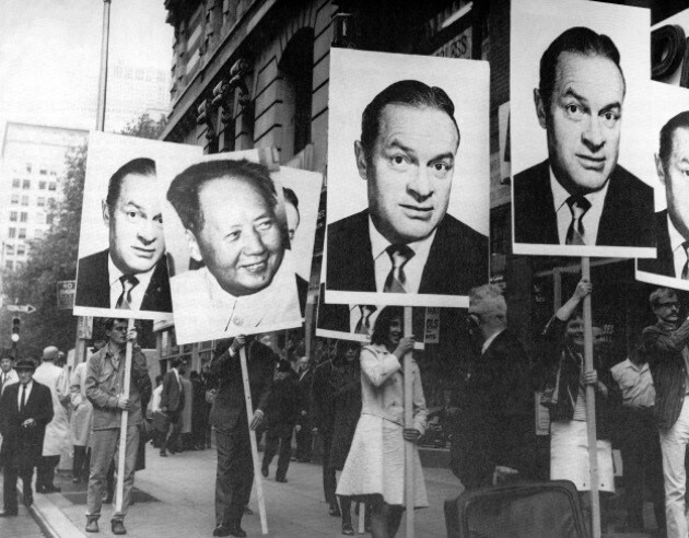 People in a street are holding large posters with black-and-white photographs of faces on them. The background includes a multi-story building and more people walking. This is an AI generated description. To give feedback, email ai@moma.org.