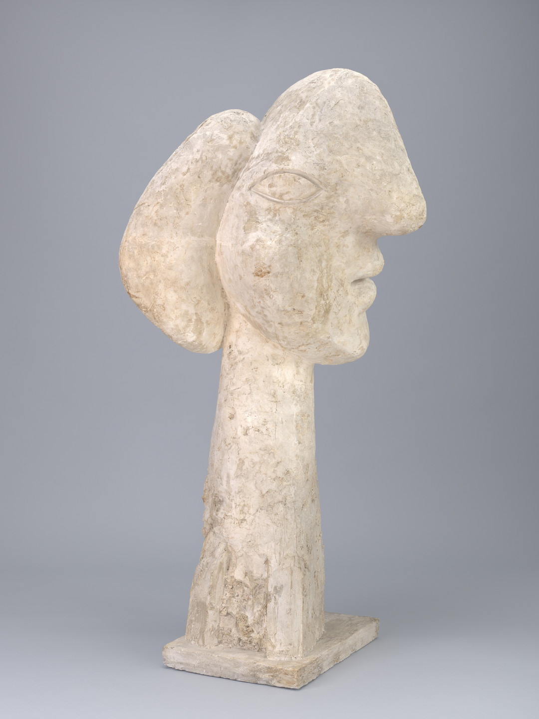 A stone sculpture depicting a stylized human head with elongated features, set on a rectangular base. The sculpture is predominantly off-white with a rough texture, set against a plain gray background. This is an AI generated description. To give feedback, email ai@moma.org.