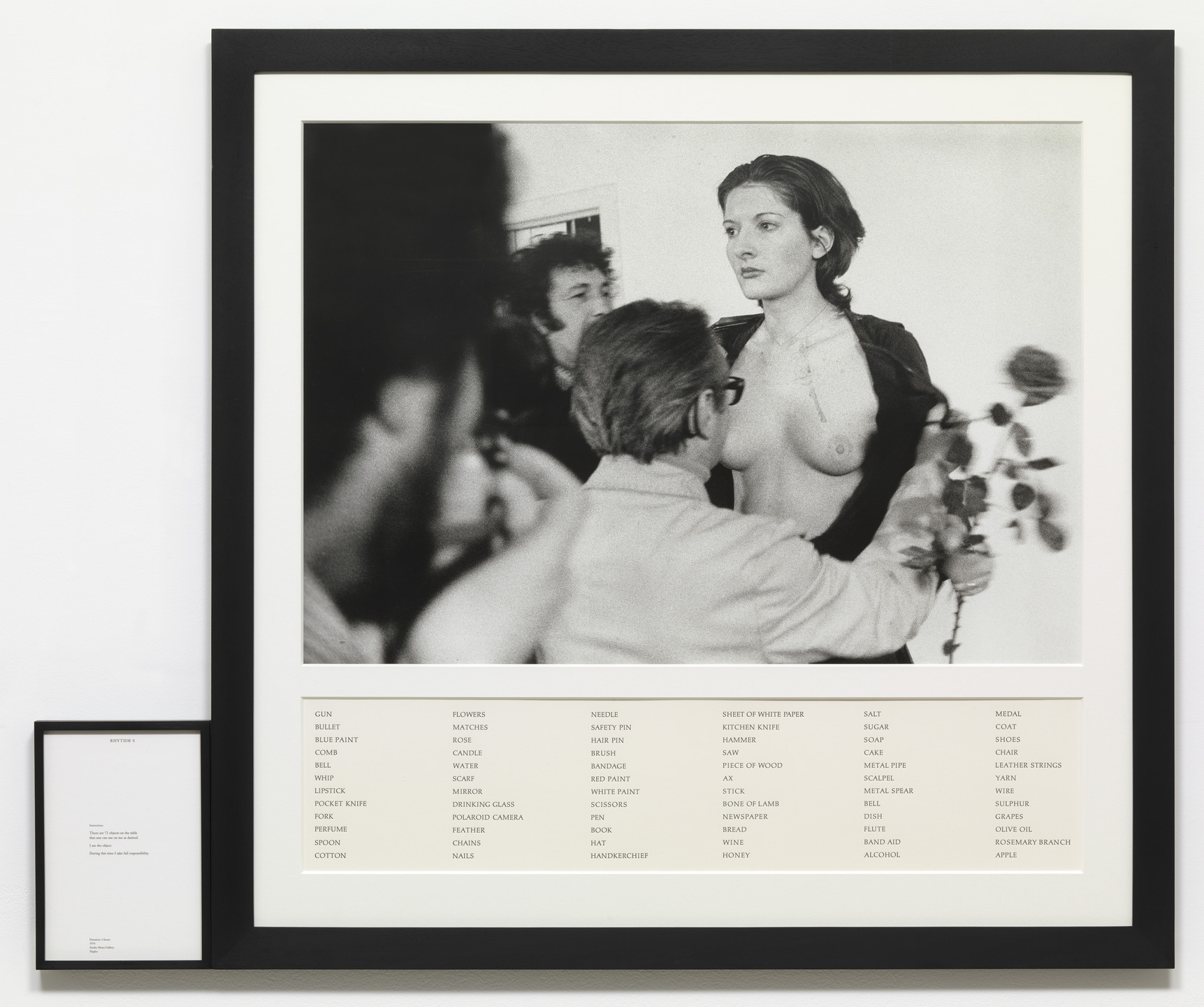 A black-and-white photo shows a person standing topless while others interact closely around them. Below the photo, there is a list of items including &#34;gun,&#34; &#34;rose,&#34; and &#34;wine&#34; in black text on a white background. This is an AI generated description. To give feedback, email ai@moma.org.
