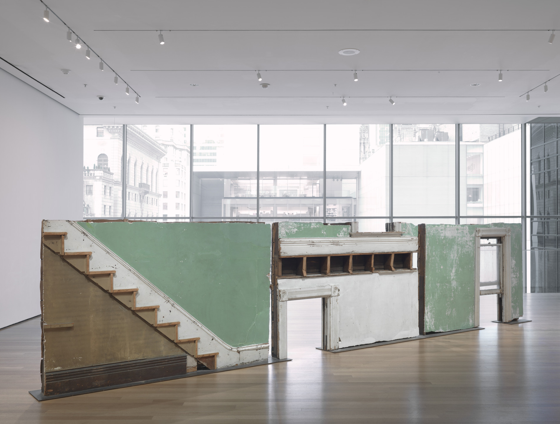 The image shows a large indoor sculpture made of cut sections of home's interior. The walls are painted green, the moulding is white, and the stairs are brown. This sculpture is positioned in a brightly lit gallery. Large windows in the background reveal an urban view. This is an AI generated description. To give feedback, email ai@moma.org.