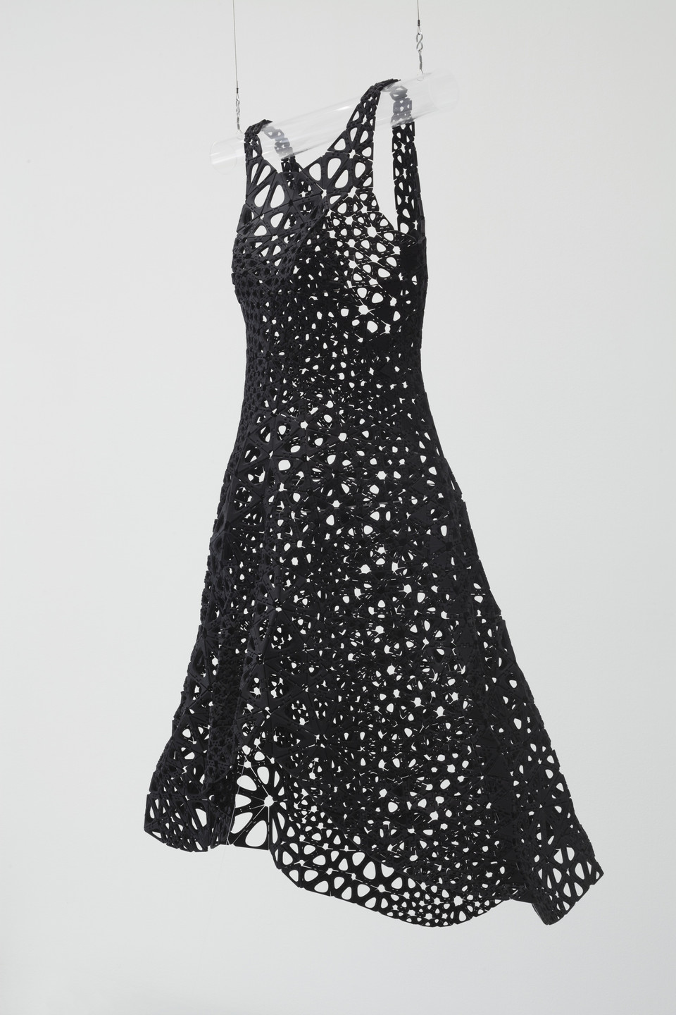A black dress with numerous circular and irregular cutouts hangs suspended on a clear hanger against a plain white background. The dress flares out slightly at the bottom. This is an AI generated description. To give feedback, email ai@moma.org.