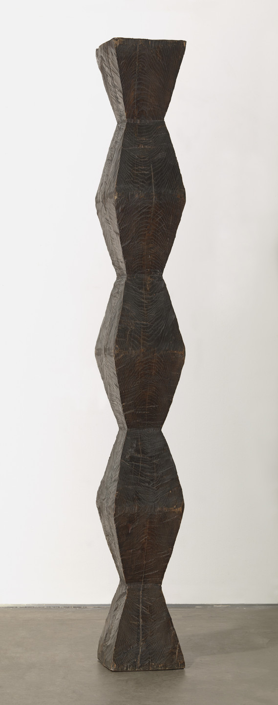 A tall, slender, sculpture made of stacked, uneven geometric shapes in deep brown and black hues with visible wood grain textures. The sculpture stands upright on a gray floor against a white background. This is an AI generated description. To give feedback, email ai@moma.org.