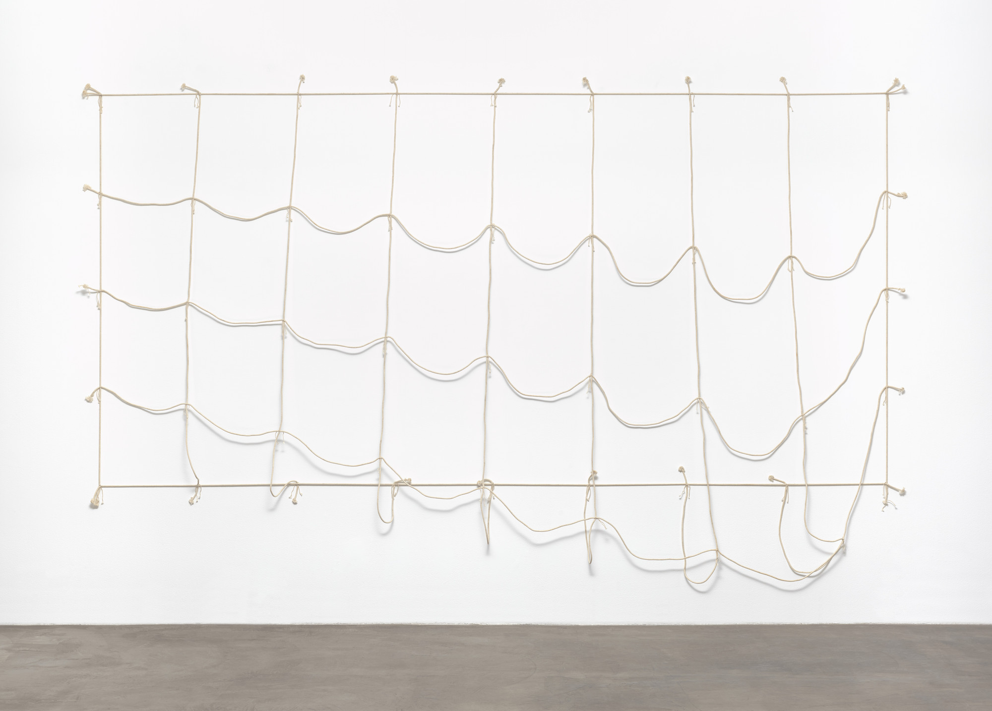 A series of wavy beige ropes are strung in a loose grid pattern against a white wall. The bottom edges of some ropes dangle freely above the gray floor. This is an AI generated description. To give feedback, email ai@moma.org.