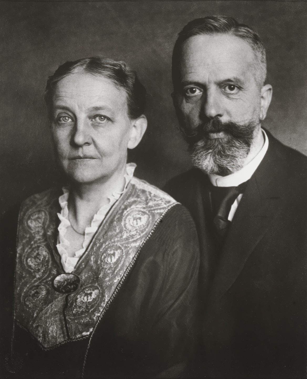 An older person with a mustache and beard, wearing a dark suit, stands next to another older person in a patterned clothing with lace collar, against a dark background. The image is in black and white. This is an AI generated description. To give feedback, email ai@moma.org.
