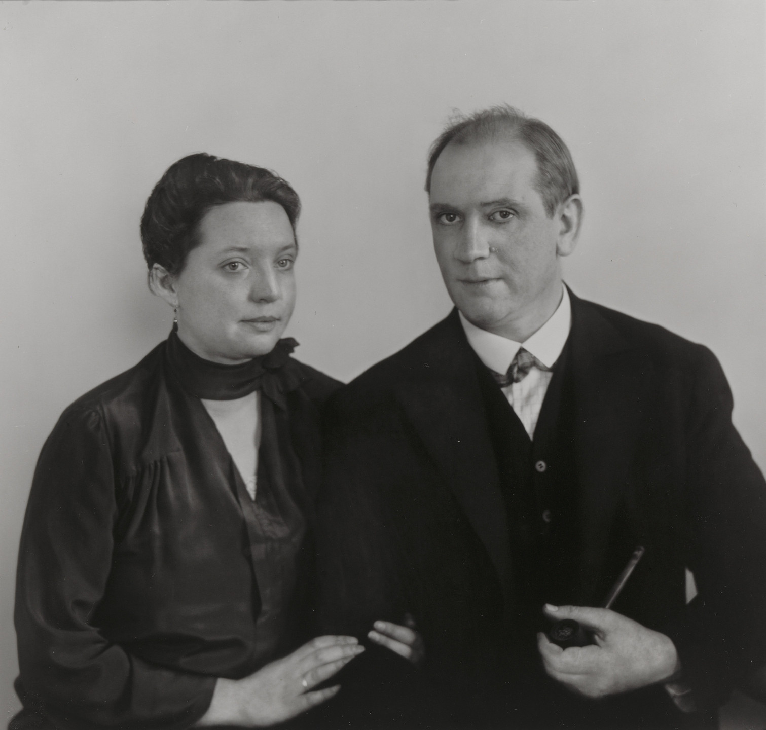 A black-and-white photograph shows two people sitting side by side, both wearing dark clothing. One person holds a pipe and has a calm expression, while the other has their hand resting on the first person&#39;s arm. This is an AI generated description. To give feedback, email ai@moma.org.