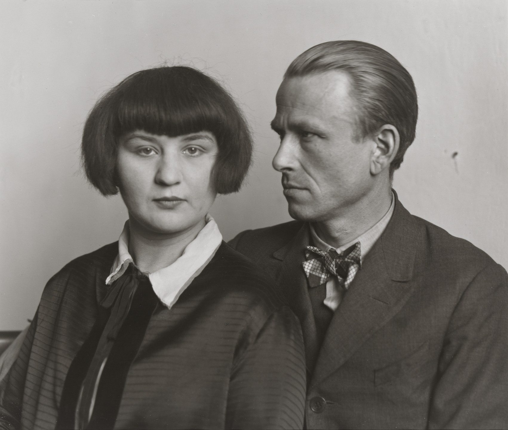 A black-and-white photo shows two individuals in close proximity. The person on the left has a straight bob haircut and a collared garment, while the person on the right, wearing a bow tie and suit, looks towards them. This is an AI generated description. To give feedback, email ai@moma.org.
