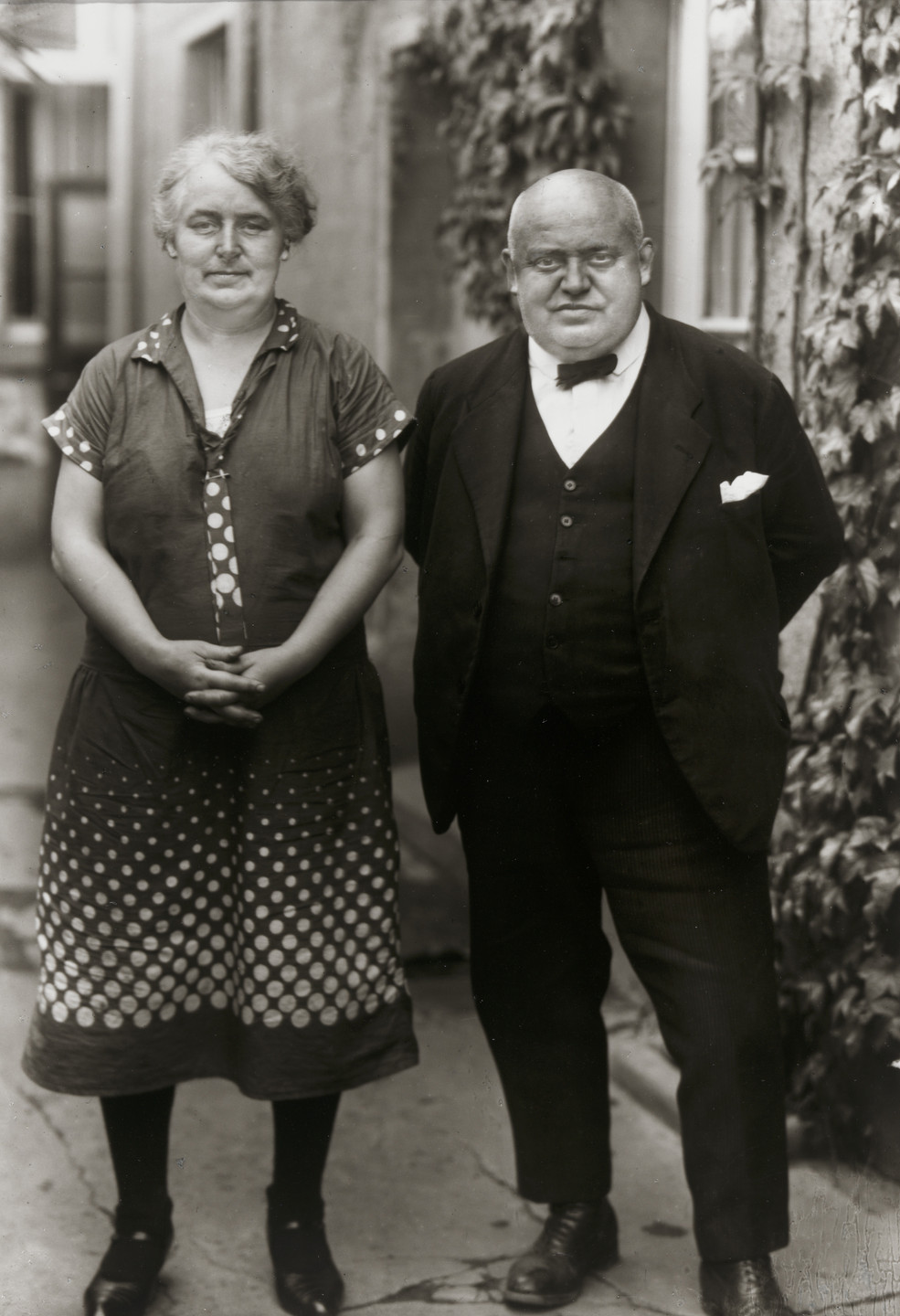 Two people are standing side by side; the person on the left is wearing a polka-dot dress and the person on the right is in a suit with a bow tie. The background shows an outdoor area with ivy-covered walls. This is an AI generated description. To give feedback, email ai@moma.org.