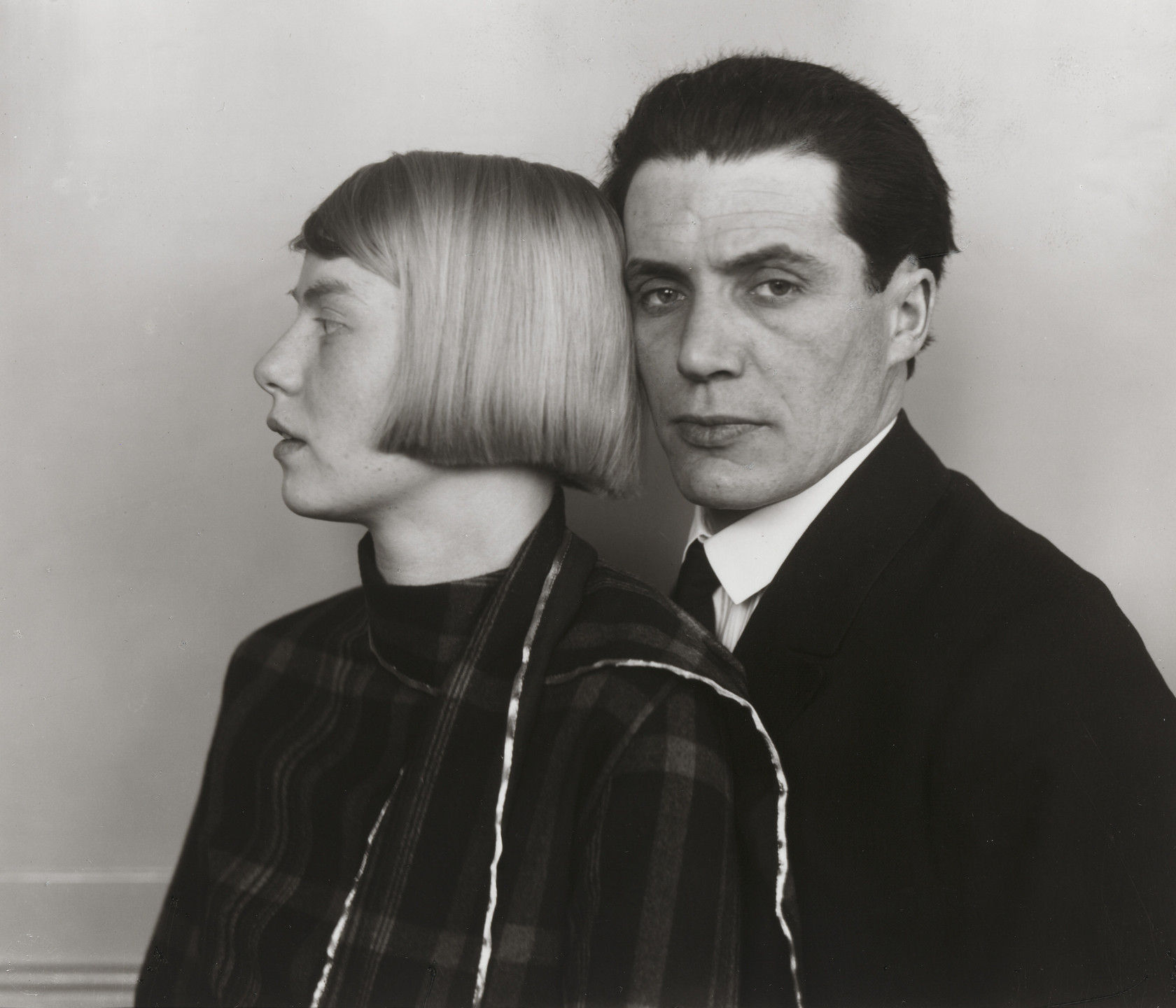 Two individuals are posed against a plain background; one faces left in profile with short, straight hair, and the other faces forward with dark, combed-back hair. The image is black and white, with the person in profile wearing a patterned garment. This is an AI generated description. To give feedback, email ai@moma.org.