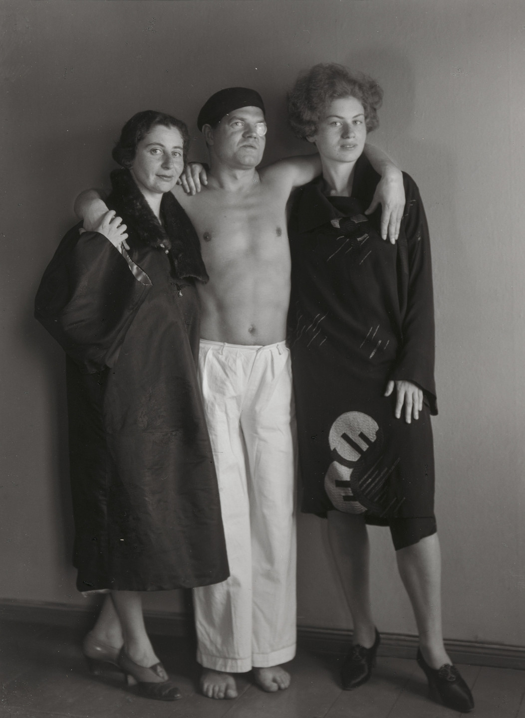 A shirtless person wearing white pants and a beret stands between two people draped in dark coats. The three individuals are standing close together against a plain background. This is an AI generated description. To give feedback, email ai@moma.org.