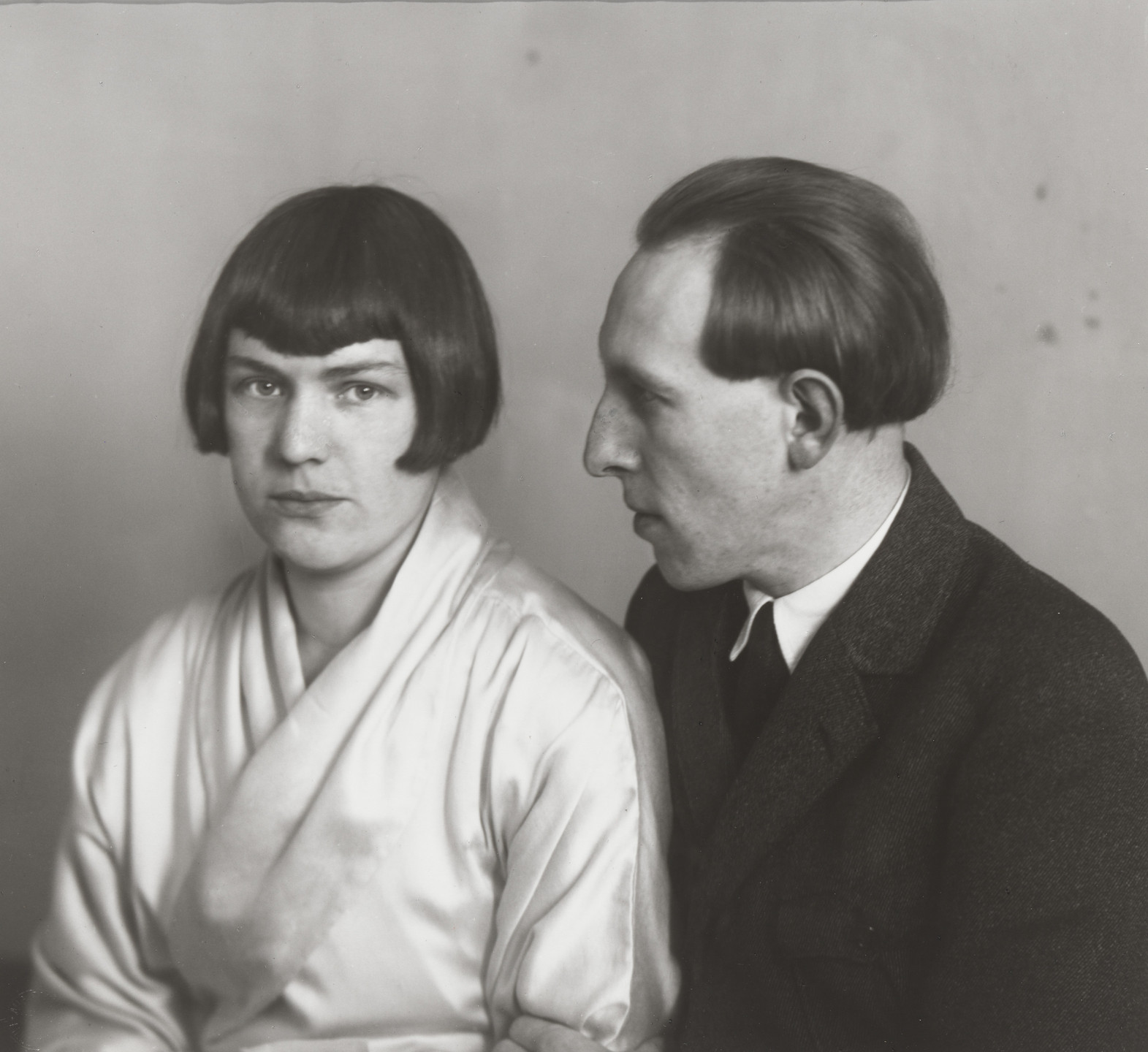 Two people are captured in a black-and-white photograph. One person with chin-length dark hair looks at the camera, wearing white, while the other person faces them with short hair, dressed in a dark blazer and tie. This is an AI generated description. To give feedback, email ai@moma.org.