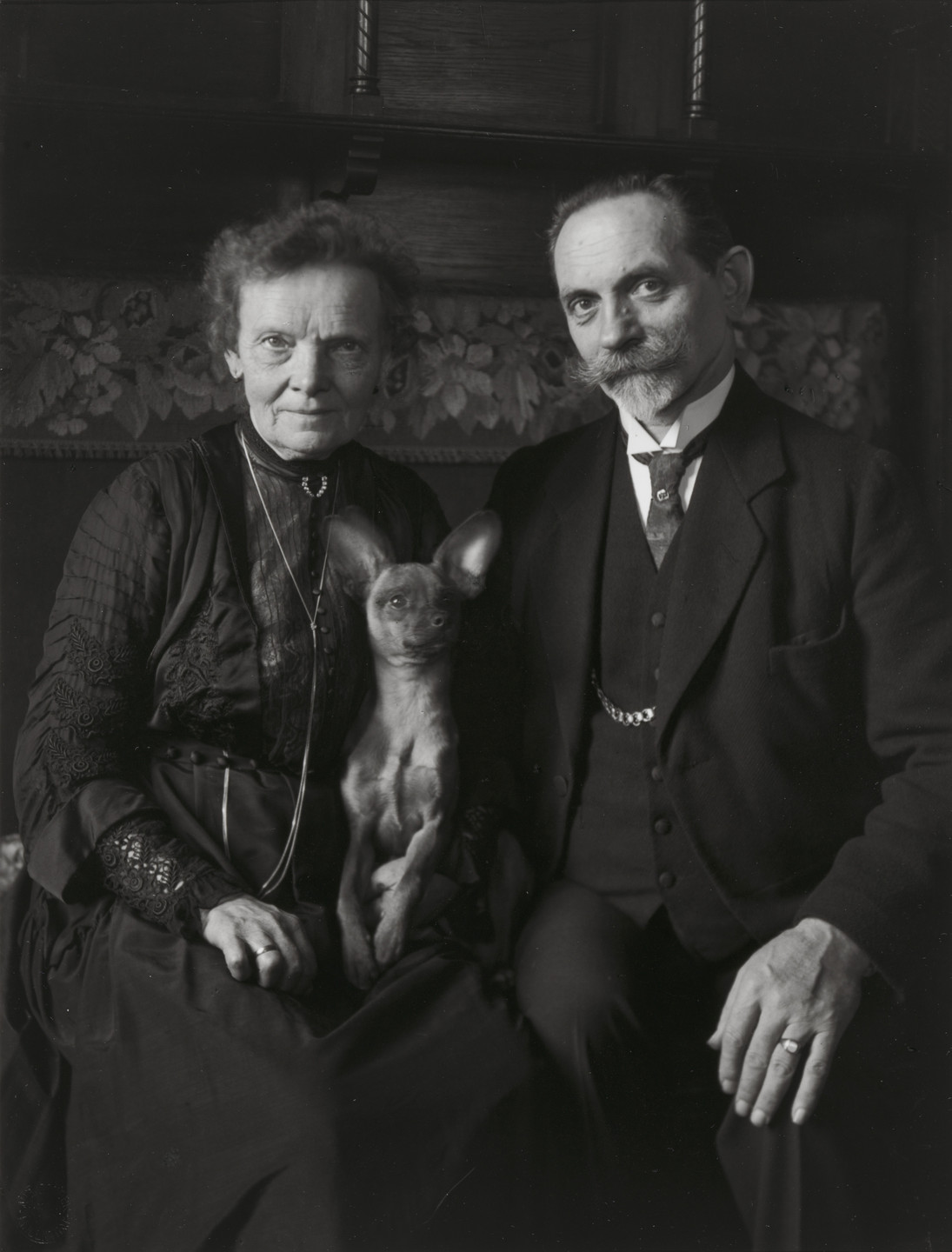 An older couple, dressed in dark, formal clothing, sit closely together with a small dog on their lap. The background features a floral pattern and dark wooden furniture. This is an AI generated description. To give feedback, email ai@moma.org.