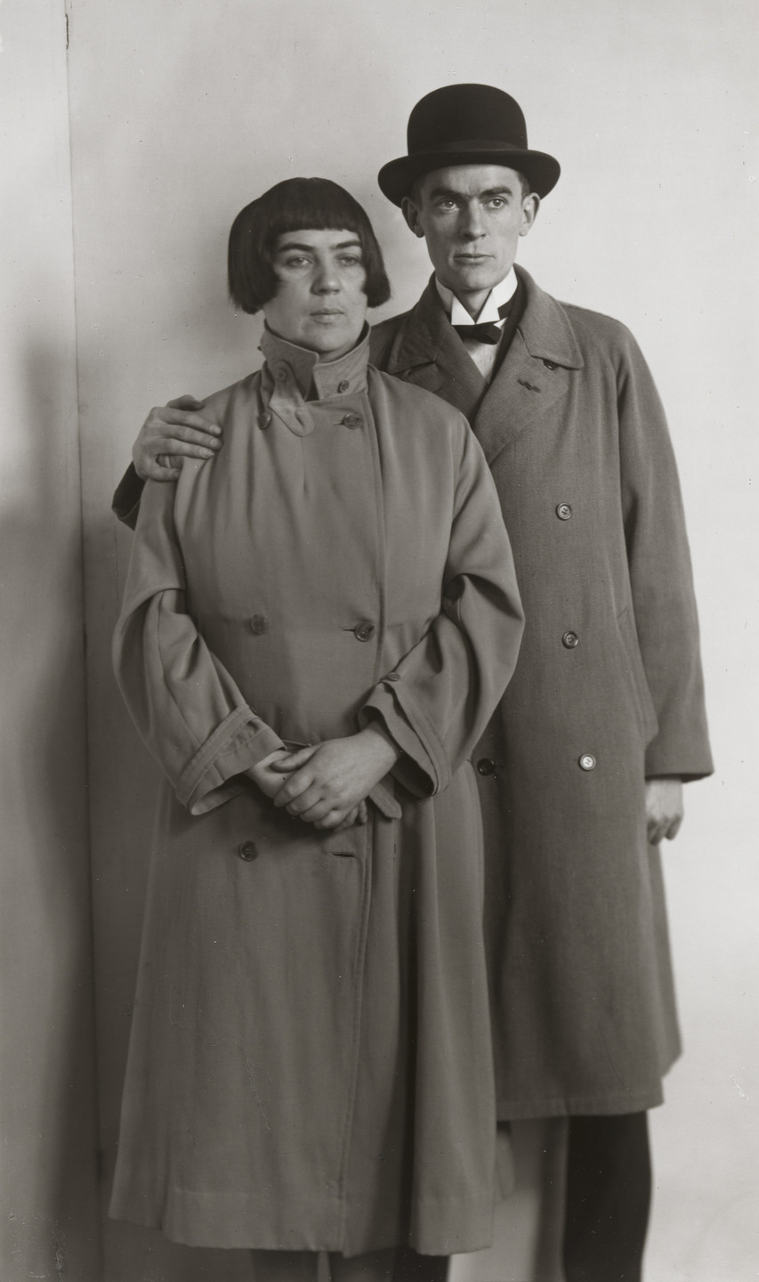 Two people in long, dark coats stand side by side against a plain background. One has a hand on the other&#39;s shoulder, while the other has hands clasped in front. This is an AI generated description. To give feedback, email ai@moma.org.