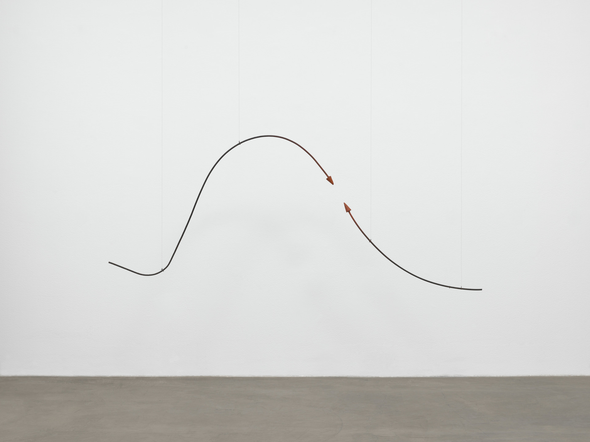 A curving black metal rod with two red arrowheads is suspended by thin wires against a plain white background above a gray floor. The rod forms a smooth wave shape from left to right. This is an AI generated description. To give feedback, email ai@moma.org.