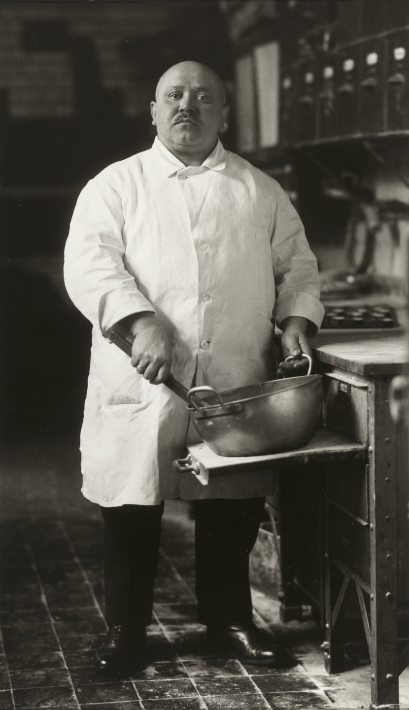 A person in a white coat holds a large pot in a kitchen setting. The floor tiles are dark, and a row of dials or gauges is visible in the background. This is an AI generated description. To give feedback, email ai@moma.org.