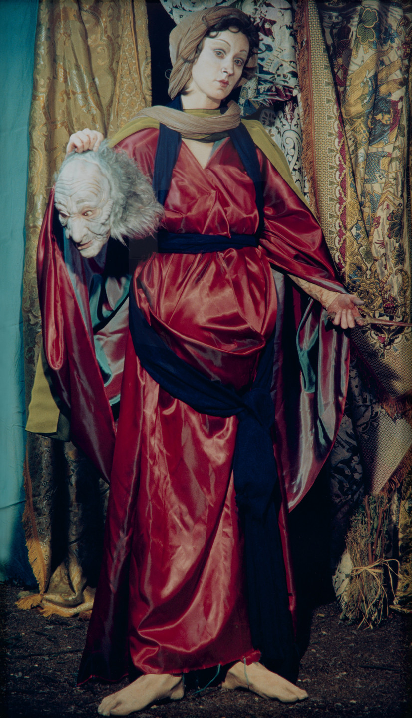The image shows a person in a red robe with a blue sash standing barefoot on a dark surface, holding a severed head with gray hair. The background features ornate, multicolored drapes. This is an AI generated description. To give feedback, email ai@moma.org.