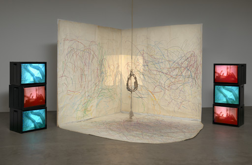 A room with off-white walls and floor has colorful squiggly lines in various shades. Six black TV screens, stacked in two columns, display blue and red images. A rope with a silver harness hangs from the ceiling. This is an AI generated description. To give feedback, email ai@moma.org.