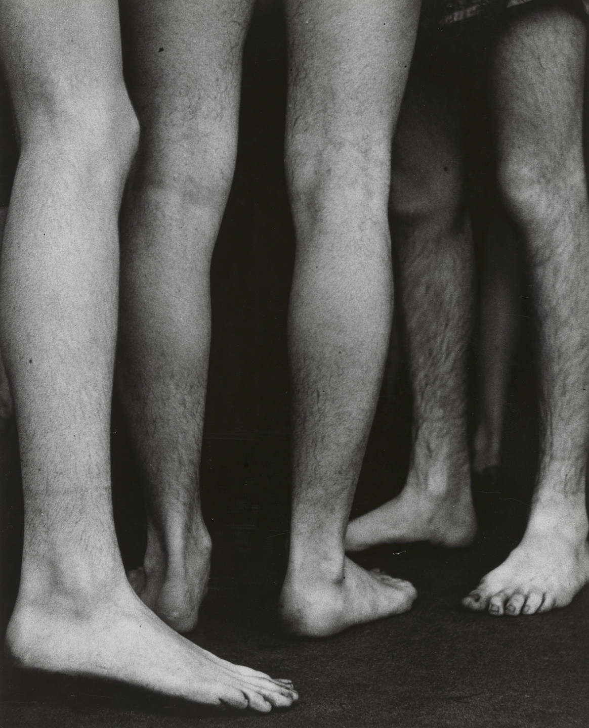 The image shows three pairs of legs and bare feet of individuals standing close together on a dark surface. The photograph is in black and white, highlighting various shadows and textures. This is an AI generated description. To give feedback, email ai@moma.org.
