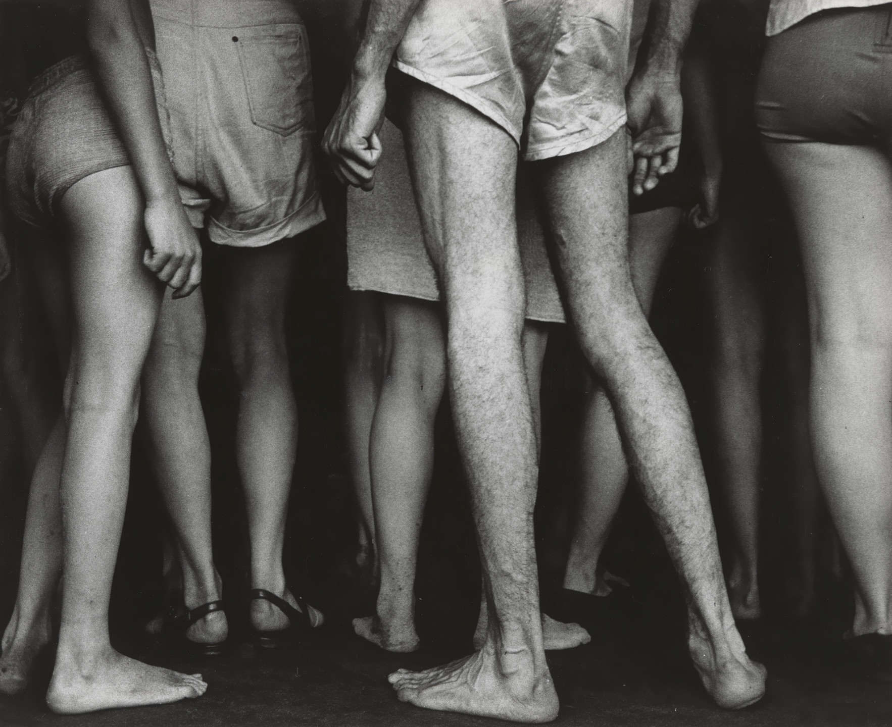 Black and white photo of several people standing close together, showcasing only their legs and lower bodies wearing shorts. The image focuses on the bare feet and calves of the group. This is an AI generated description. To give feedback, email ai@moma.org.