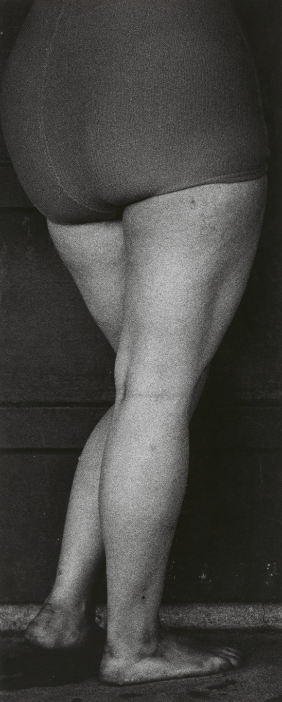A grayscale photo shows the lower body of a person standing barefoot on a rough surface. The person is wearing shorts and their legs are crossed at the ankles. This is an AI generated description. To give feedback, email ai@moma.org.