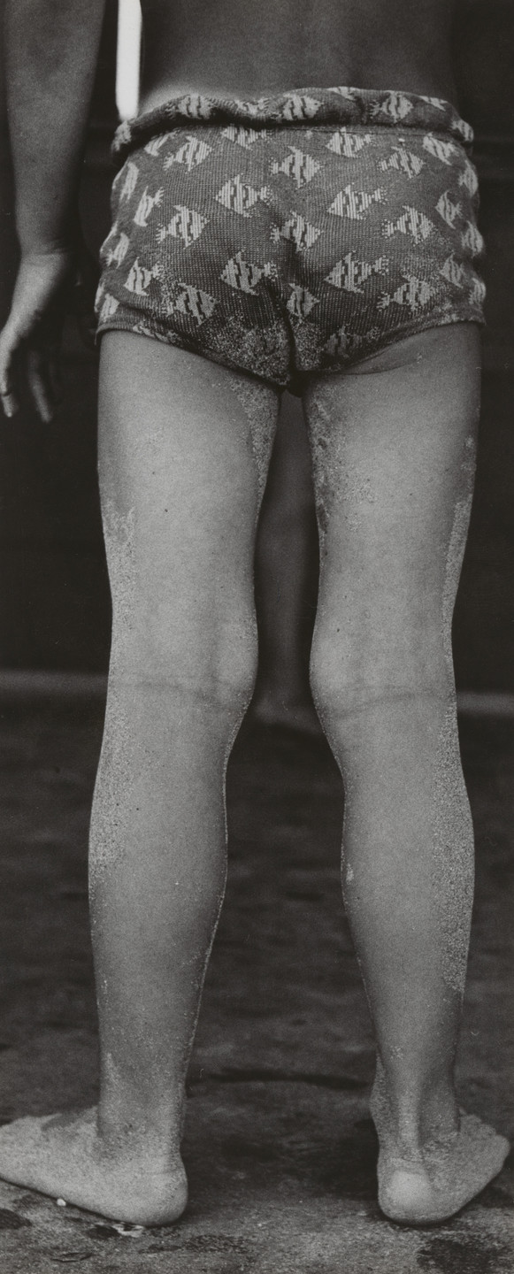 A person stands with their back facing the camera, wearing patterned shorts with many small shapes. The person&#39;s legs and feet are covered in sand, and the image is in black and white. This is an AI generated description. To give feedback, email ai@moma.org.