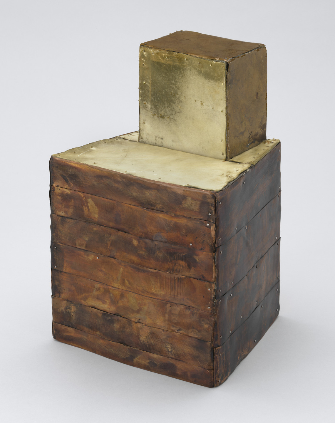 The image features a rectangular, vertically oriented, metallic sculpture with two stacked sections. The bottom part is dark brown with visible seams, and the top part is a lighter, tarnished gold color. This is an AI generated description. To give feedback, email ai@moma.org.