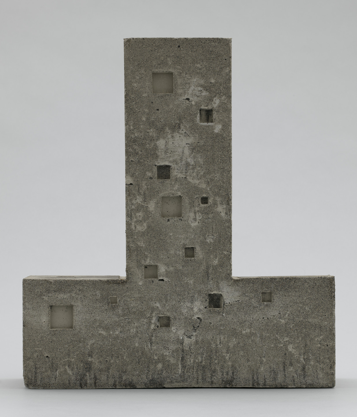 A concrete structure shaped like an upside-down &#34;T&#34; with nine small square cutouts of varying sizes scattered across its rough-textured surface on a plain grey background. The structure is predominantly gray, with darker patches around some cutouts. This is an AI generated description. To give feedback, email ai@moma.org.