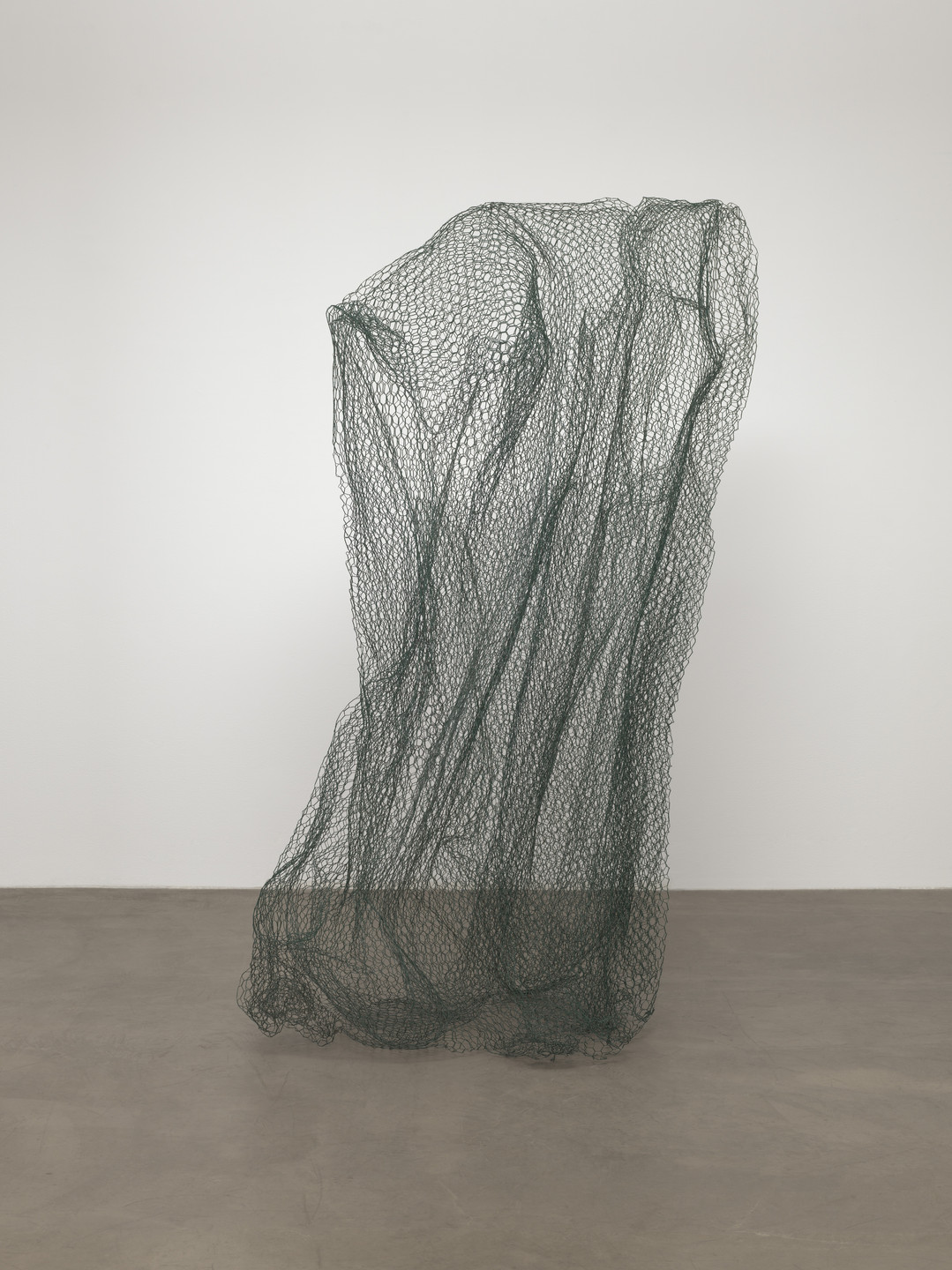 A green netting structure, vertically stretched and loosely shaped, stands on a gray concrete floor against a white background. The netting appears delicate with an intricate pattern of interlinked loops. This is an AI generated description. To give feedback, email ai@moma.org.