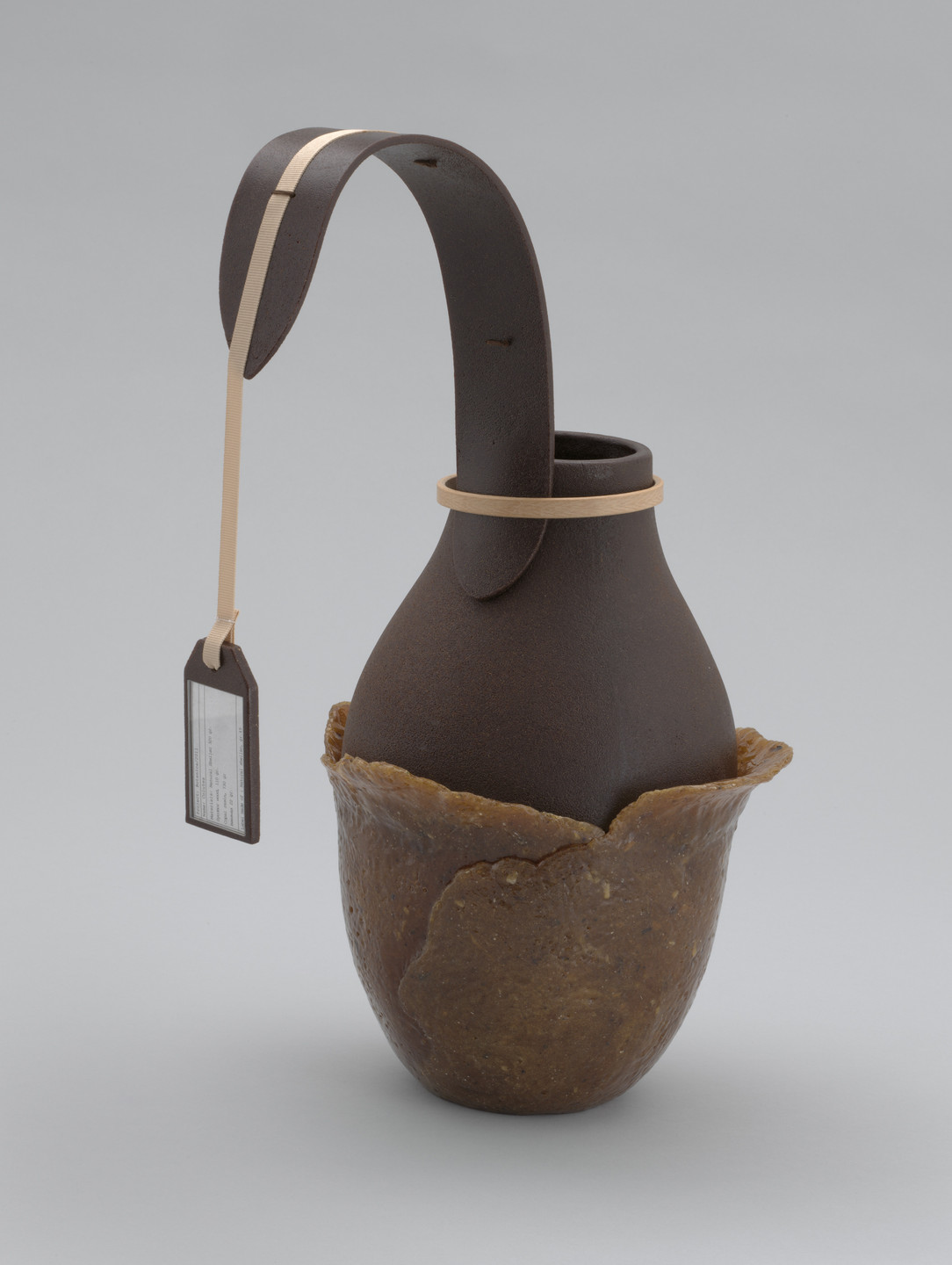 A brown vessel with a smooth, rounded body sits inside a rougher, tan-colored base, featuring a long, curved handle with a tan strap and attached tag. The background is light grey. This is an AI generated description. To give feedback, email ai@moma.org.