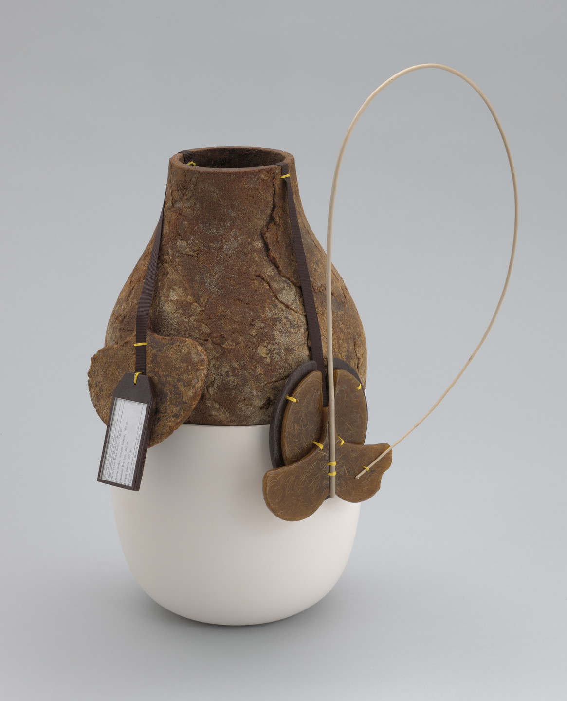 A two-toned vase with a rough, rust-colored upper section and a smooth, off-white bottom. The vase is adorned with brown, heart-shaped tags and a thin, looping handle. This is an AI generated description. To give feedback, email ai@moma.org.