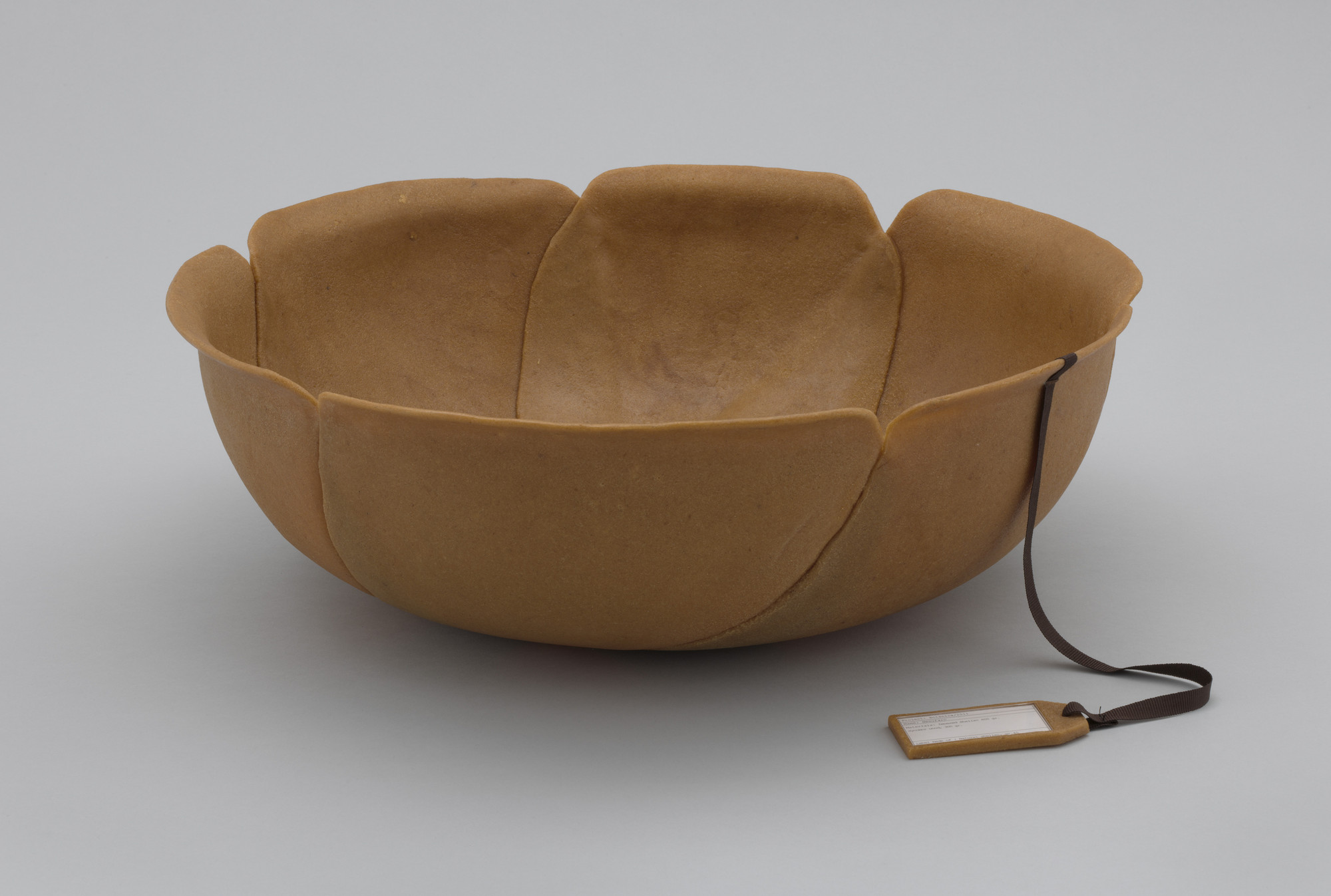 A tan-brown bowl with petal-like edges and a smooth texture sits on a light grey surface. A dark brown ribbon with a small rectangular tag is attached to the edge of the bowl. This is an AI generated description. To give feedback, email ai@moma.org.