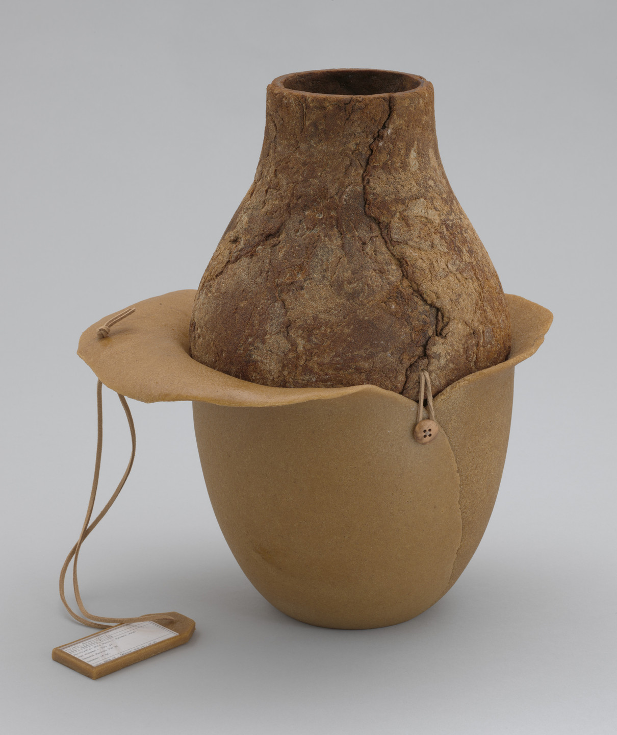 A two-toned brown vessel with a rough texture on the top half sits in a smooth, lighter brown base. It has a strap with a small tag hanging from it. This is an AI generated description. To give feedback, email ai@moma.org.