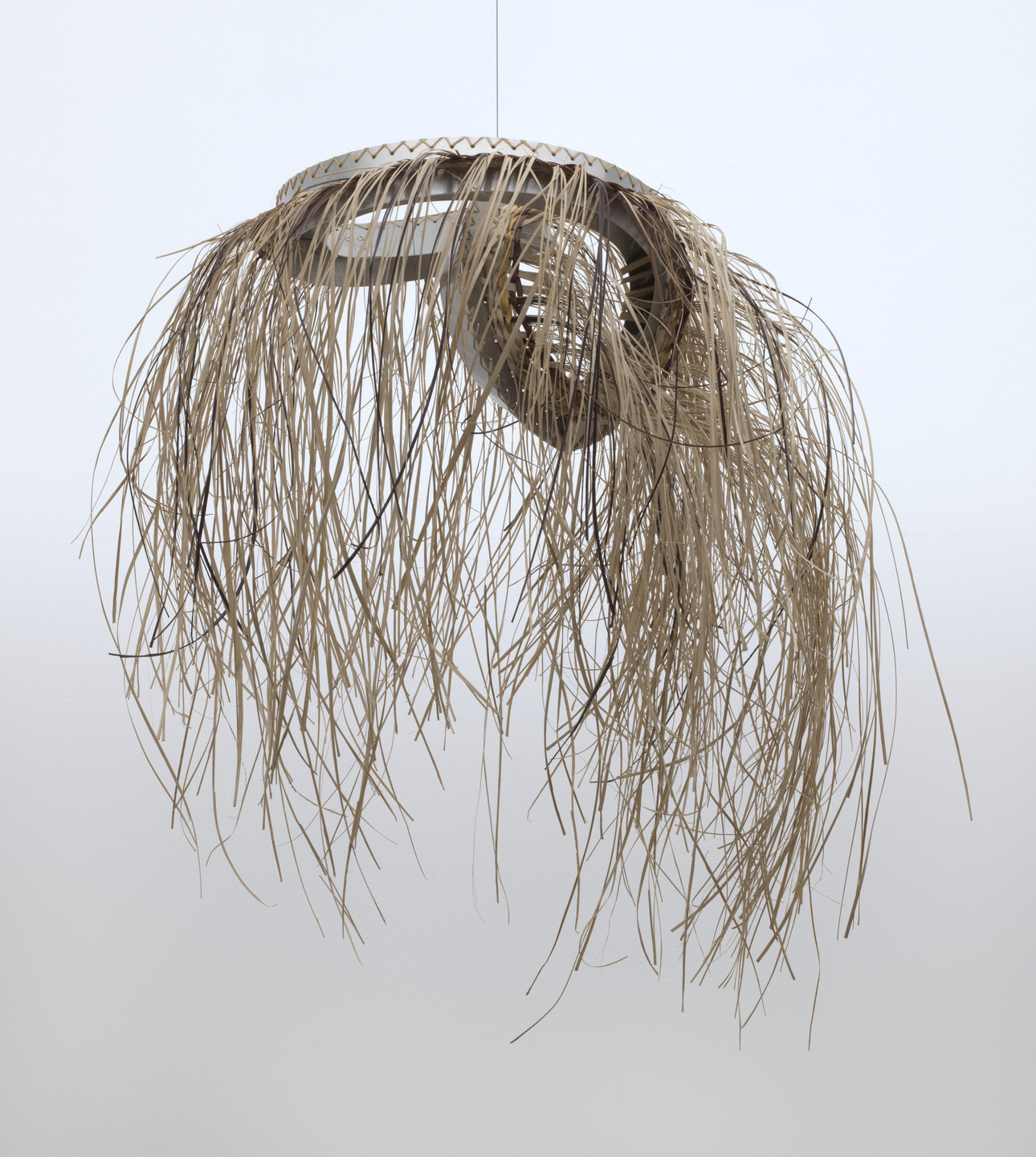 A hanging sculpture features a circular frame with long, thin, beige strands draping down from it. The background is light gray, and the overall color of the sculpture is beige. This is an AI generated description. To give feedback, email ai@moma.org.