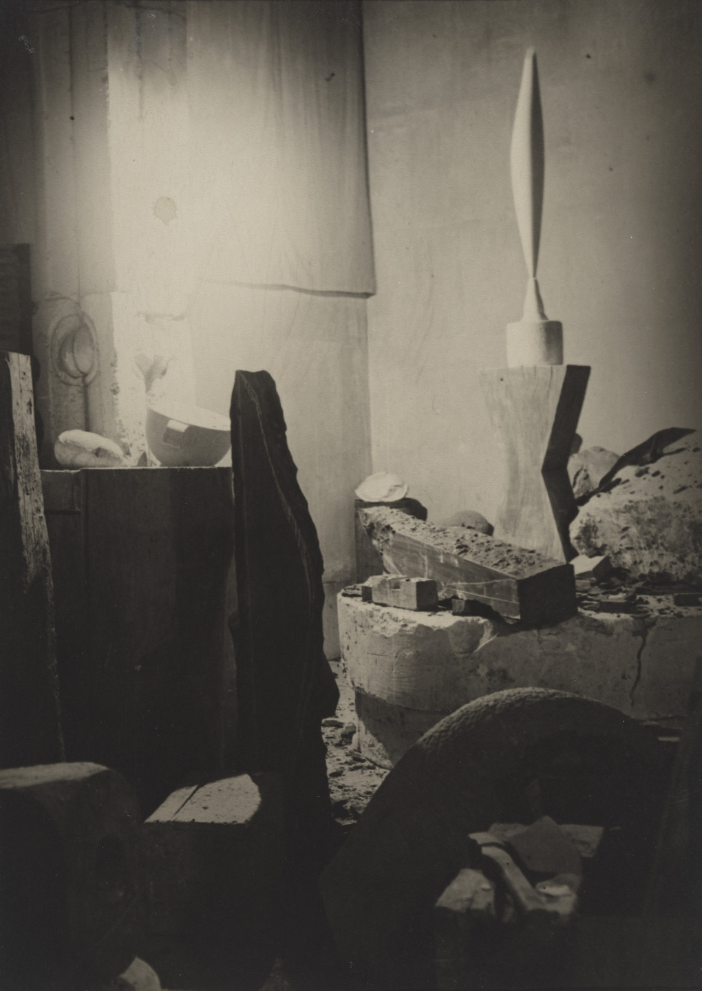 The image shows a dimly lit room with various stone-like objects and sculptures. The predominant colors are shades of gray and brown, with a tall, pointed sculpture on a stand in the background. This is an AI generated description. To give feedback, email ai@moma.org.