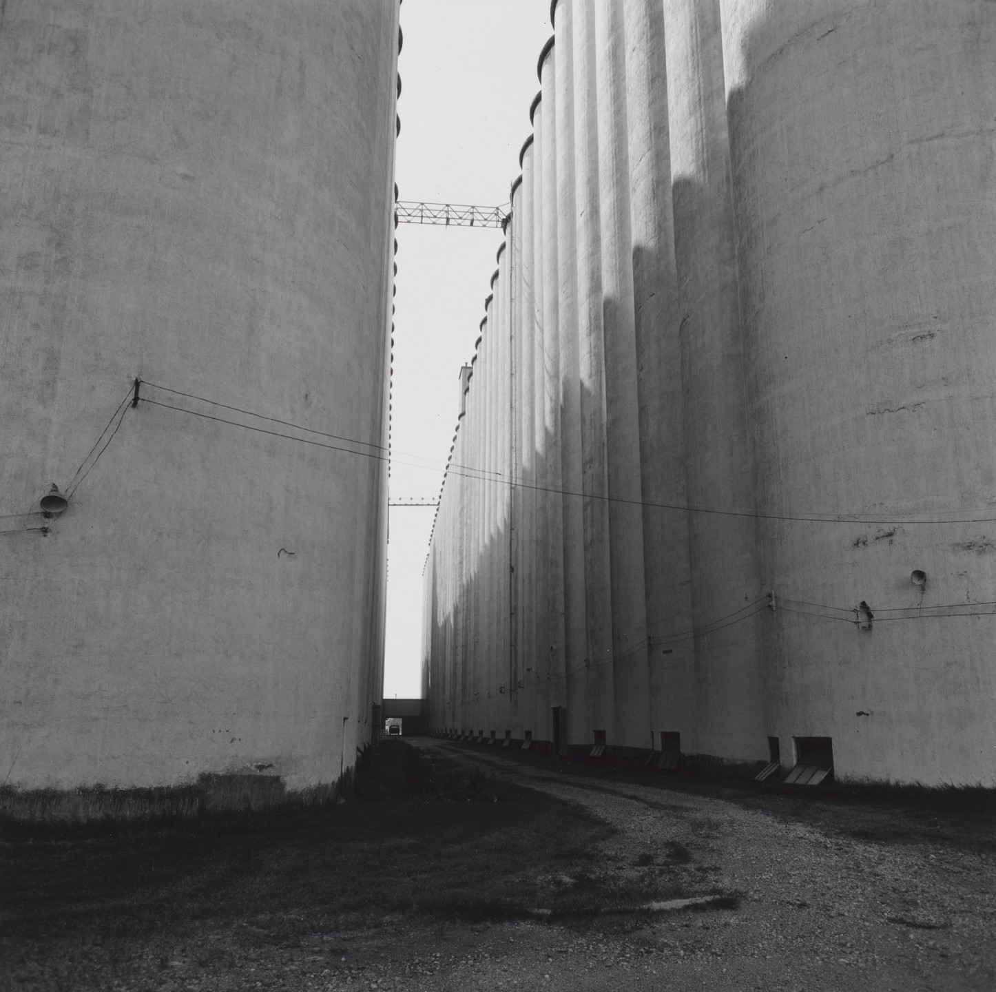 The image shows a narrow pathway bordered by very tall, cylindrical concrete structures with visible shadows. The predominant color in the image is gray. This is an AI generated description. To give feedback, email ai@moma.org.