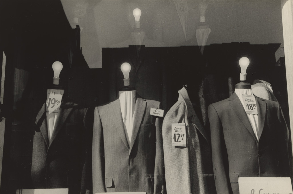 A black-and-white photograph by Lee Friedlander showing a shop window display with four mannequins wearing suits, but instead of heads, each has a glowing lightbulb. Price tags dangle from their chests. The window reflects the street outside.