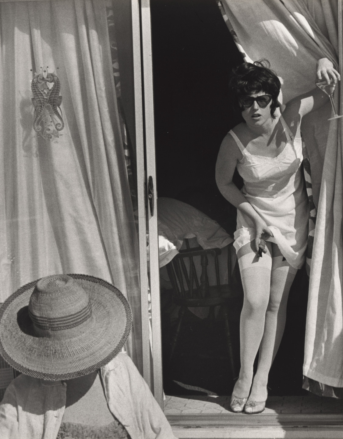 Two people are in the black and white image. One person, in stockings and holding a drink, stands in a doorway with curtains, while the other person, seated and wearing a large hat, is in the foreground. This is an AI generated description. To give feedback, email ai@moma.org.