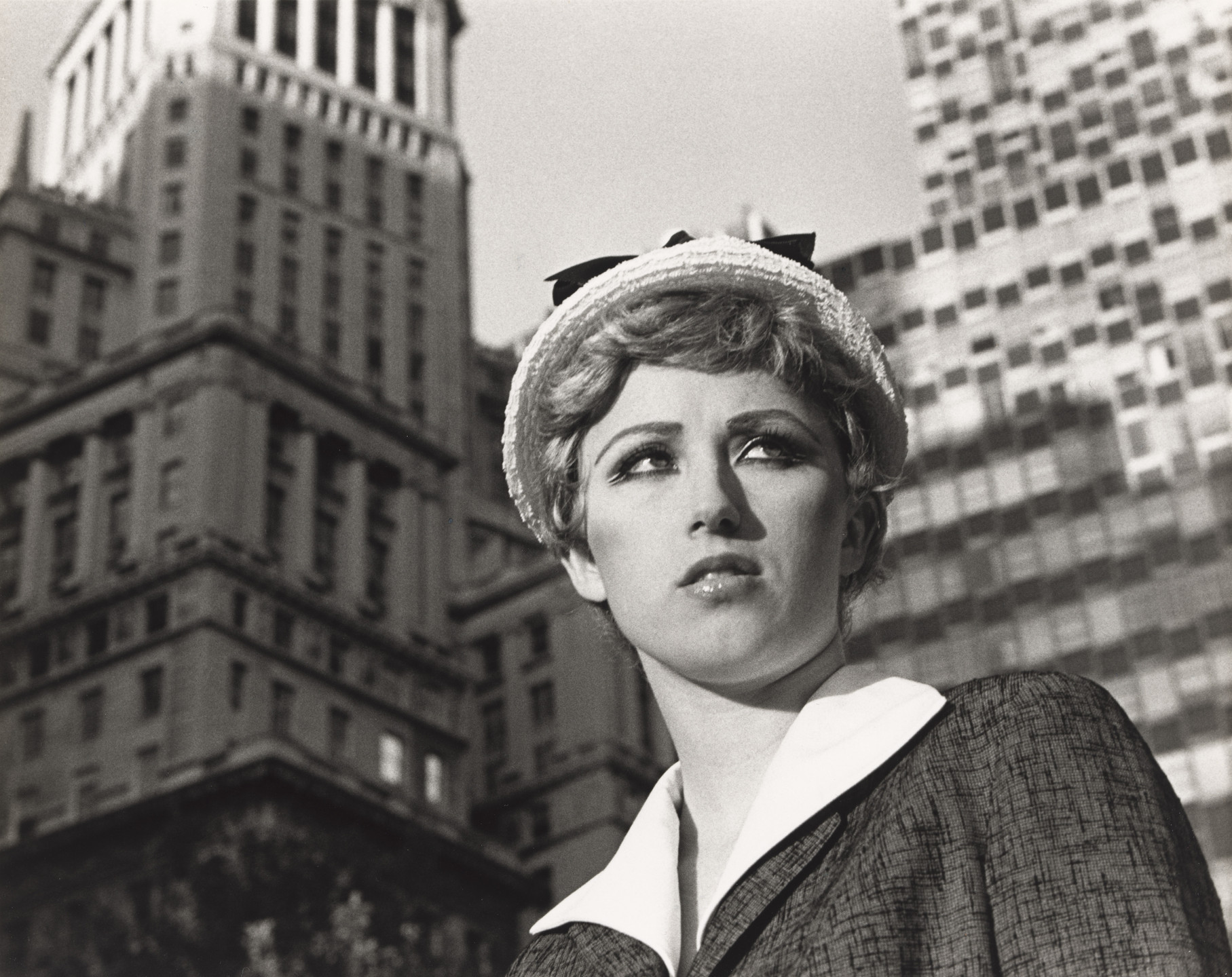 A person with short, wavy hair and a hat looks to the side with skyscrapers in the background. The image is in black and white. This is an AI generated description. To give feedback, email ai@moma.org.