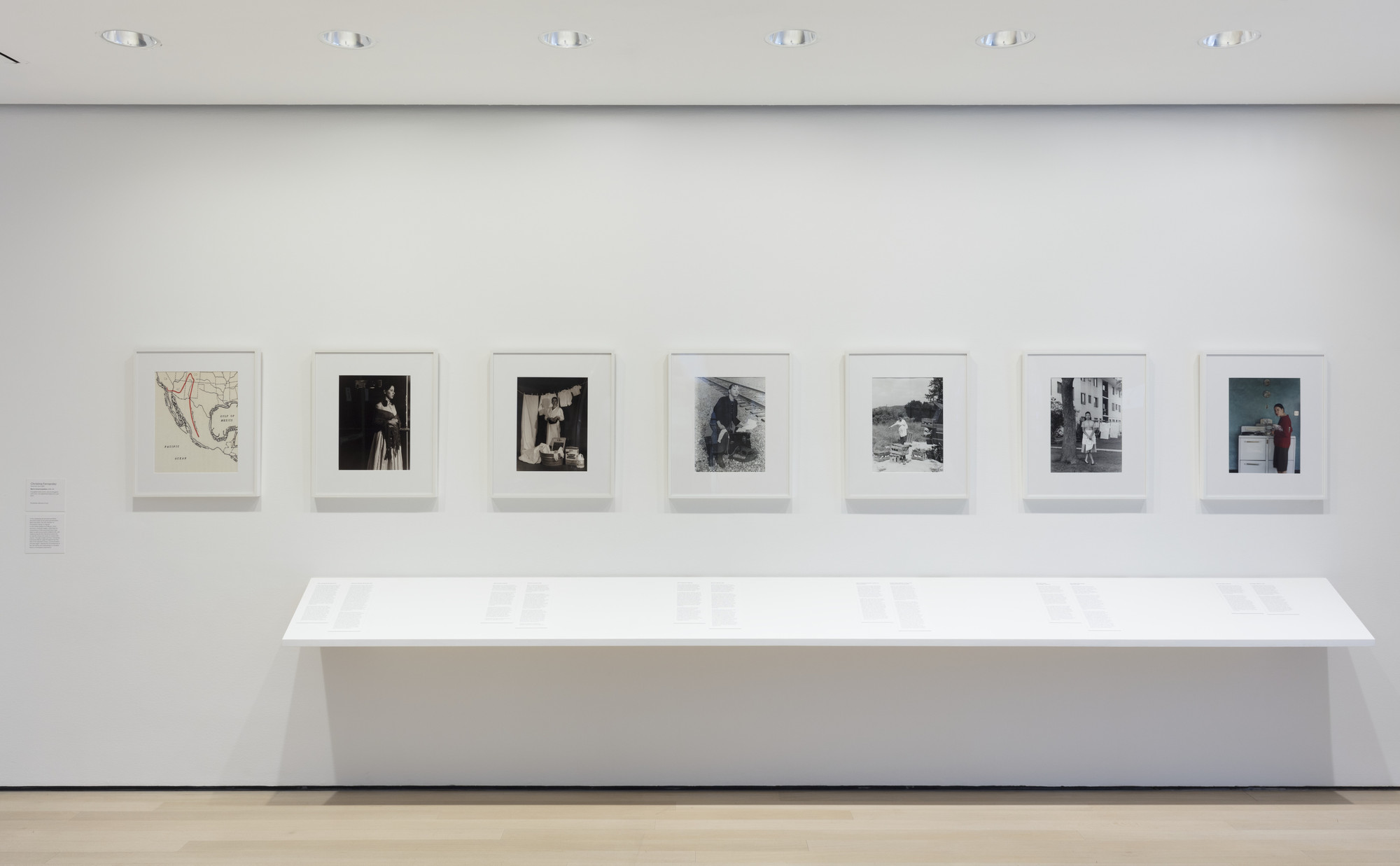 A white wall displays seven framed works, six black-and-white images and one color photograph. Below the frames is a long, white shelf with documents laid out for viewing. This is an AI generated description. To give feedback, email ai@moma.org.