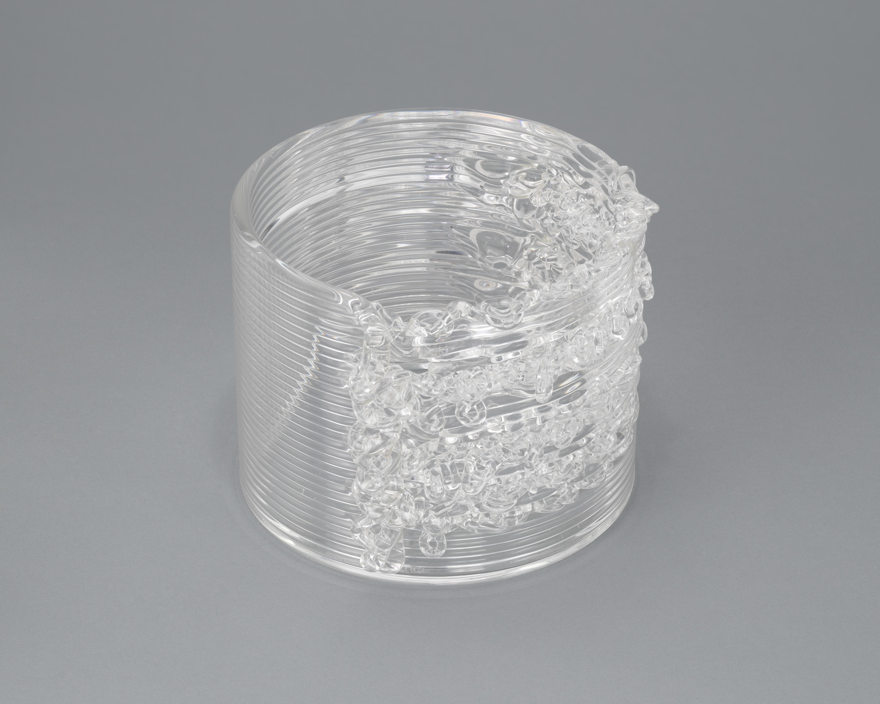 A cylindrical, clear glass sculpture with horizontal grooves and intricate, irregular patterns on one side sits on a gray background. The object appears transparent and textured, with light reflections. This is an AI generated description. To give feedback, email ai@moma.org.