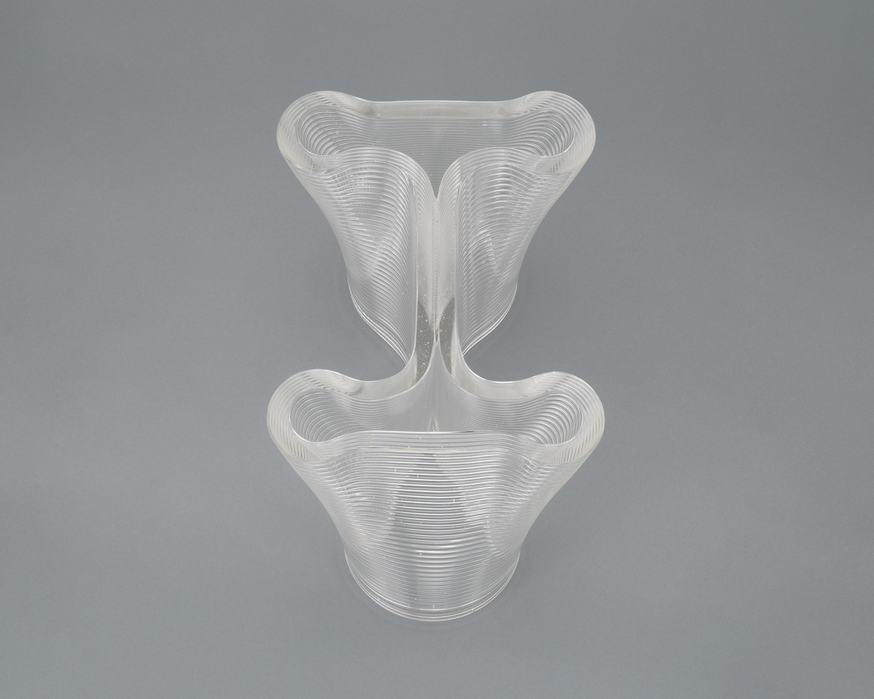 A transparent, wavy vase-like object with layered, curved ridges on a gray background. The object has a symmetrical design with deep, inward curves on both sides. This is an AI generated description. To give feedback, email ai@moma.org.