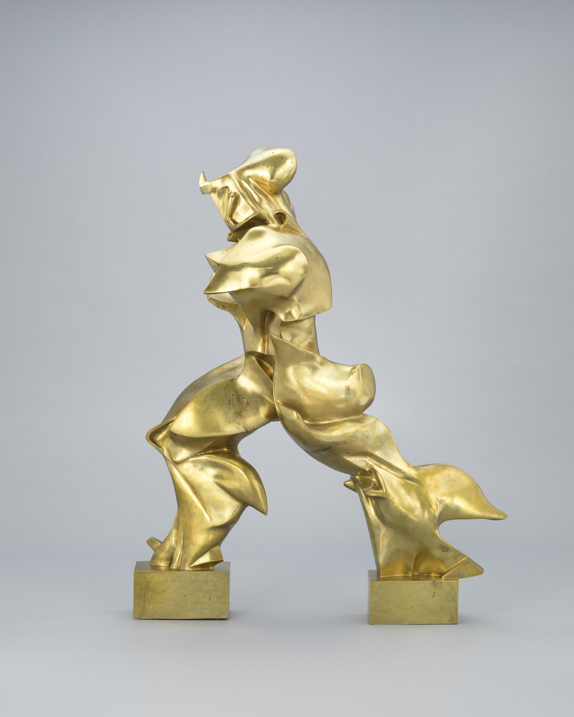 A shiny gold-colored sculpture of an abstracted human figure striding forward with its left leg. The figure appears to wear a hood and has no arms. Rippling, flame-like forms convey that the figure is in motion. Each leg is supported by a gold-colored block. This image shows the figure in profile facing left. This is an AI generated description. To give feedback, email ai@moma.org.