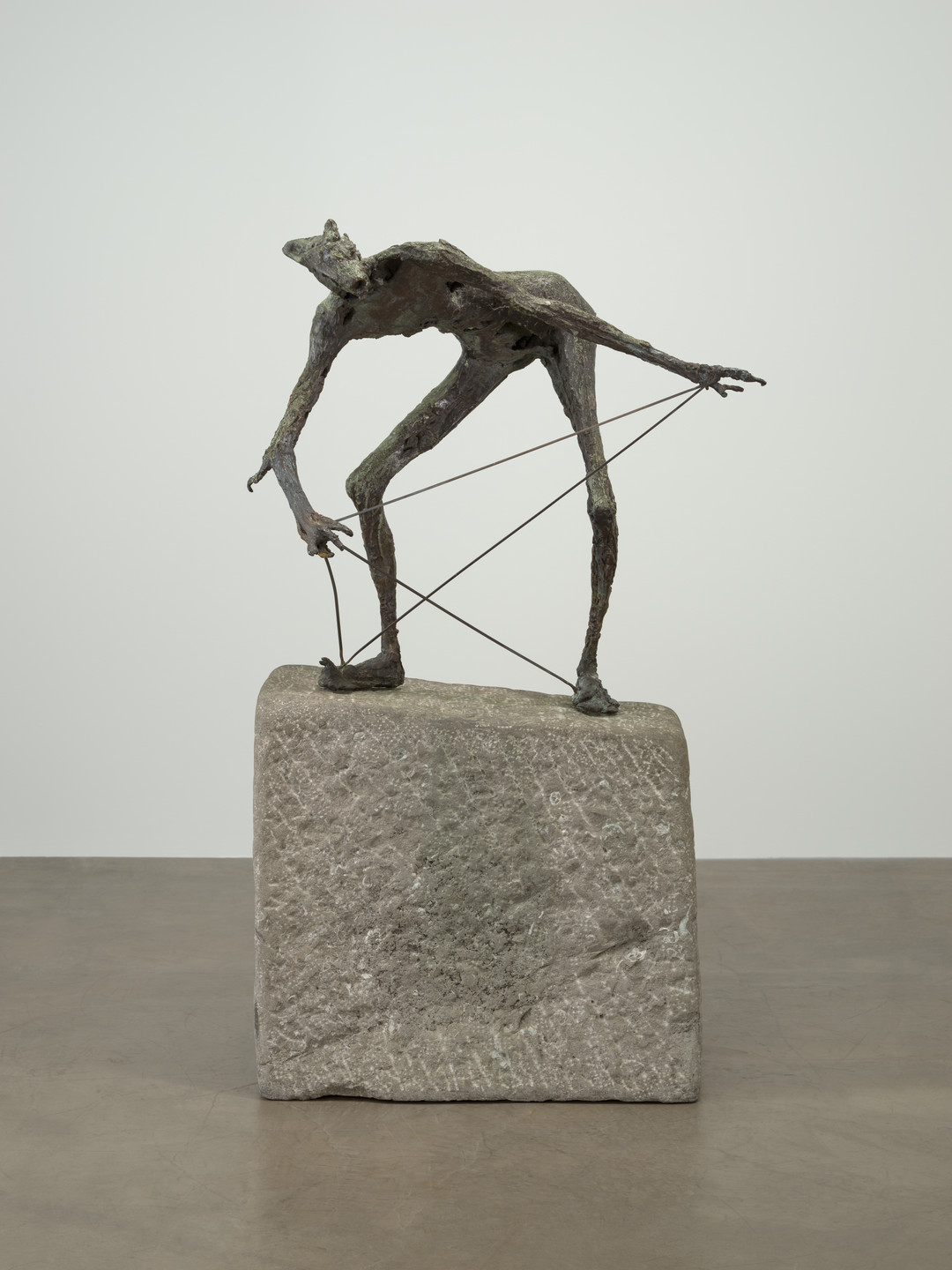 A dark bronze-colored sculpture on a rough, square stone base depicts a lanky, elongated figure with outstretched arms and a hunched posture. Thin strings are attached to its limbs, adding tension and geometric patterns. This is an AI generated description. To give feedback, email ai@moma.org.