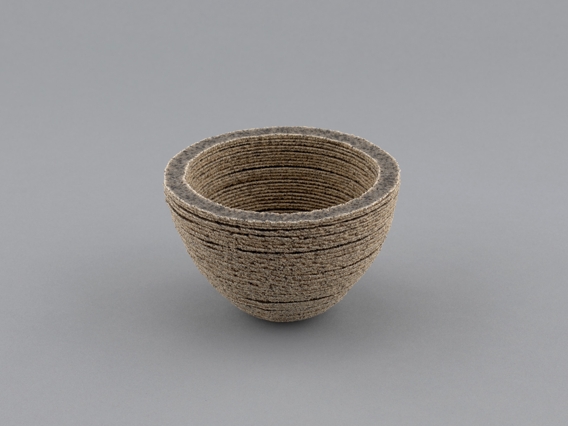 A round, textured bowl with a grayish-brown color is centered against a plain gray background. The bowl has a slightly tapered bottom and distinct horizontal lines encircling it. This is an AI generated description. To give feedback, email ai@moma.org.