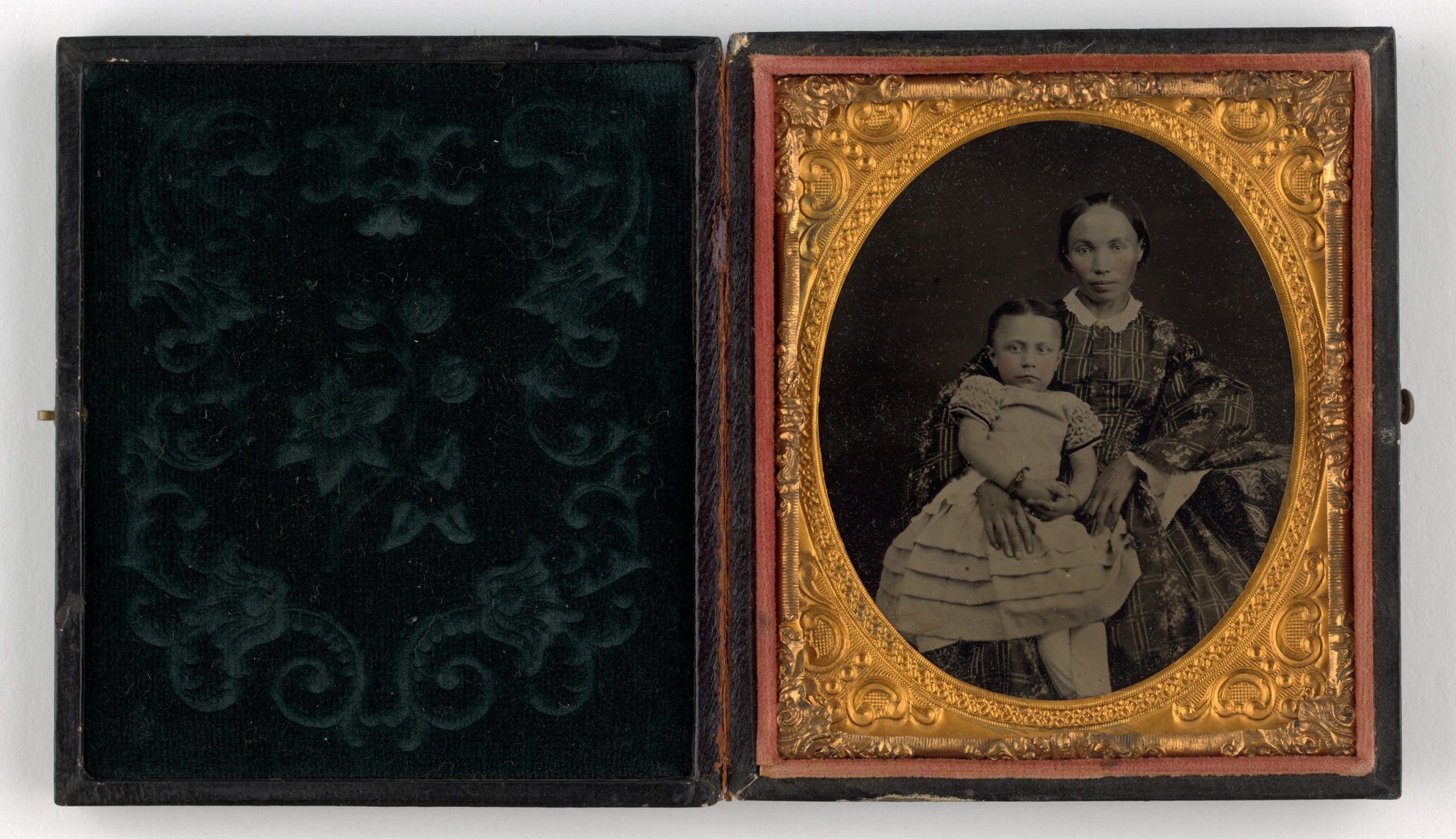 A vintage photograph shows an adult and a child seated together in a gold oval frame. They both wear patterned clothing with predominantly dark shades, in a case with intricate floral designs. This is an AI generated description. To give feedback, email ai@moma.org.