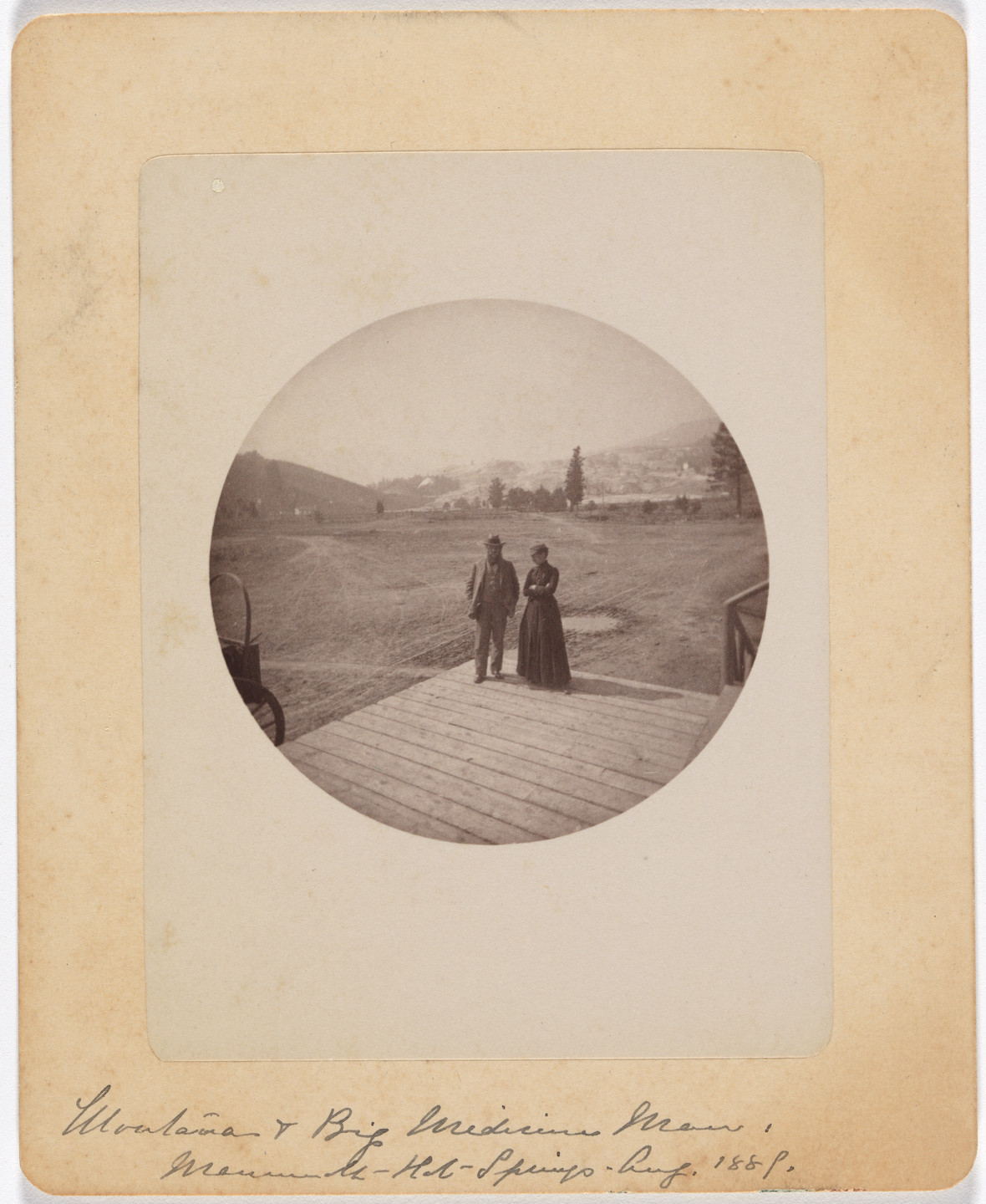 A sepia-toned photograph shows two people standing on a wooden deck with a spacious, grassy landscape and distant hills behind them. The image is oval-shaped and mounted on a tan background. This is an AI generated description. To give feedback, email ai@moma.org.