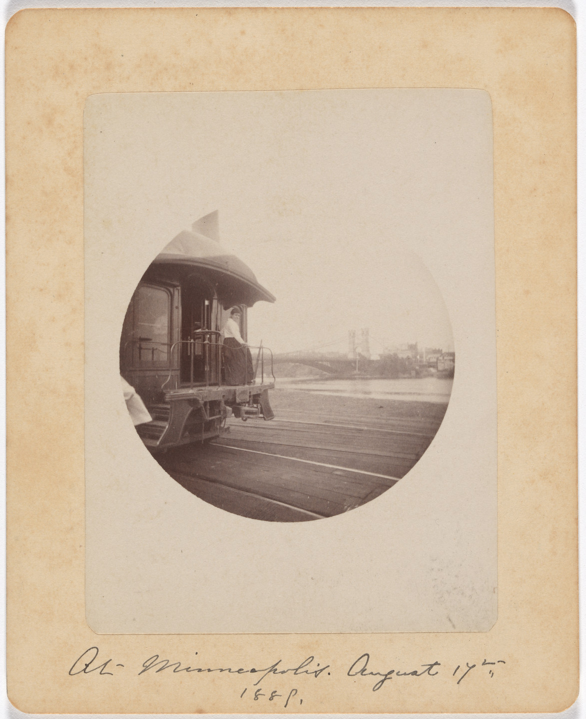 A sepia-toned photograph shows the back of a train car with a person standing on its platform. In the background, there&#39;s water and a bridge with some buildings. This is an AI generated description. To give feedback, email ai@moma.org.