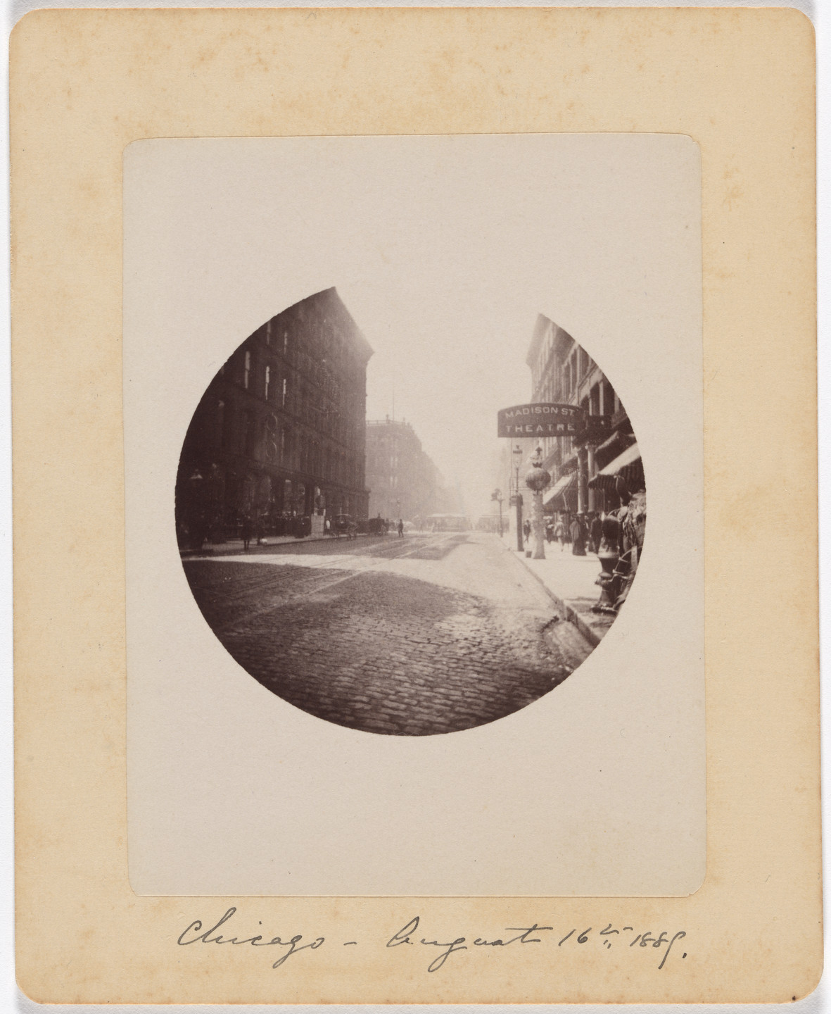 A black-and-white photograph depicts a street scene with cobblestone pavement and tall buildings on both sides. A sign for &#34;Madison St Theatre&#34; is visible on the right; people and horse-drawn carriages are present in the background. This is an AI generated description. To give feedback, email ai@moma.org.