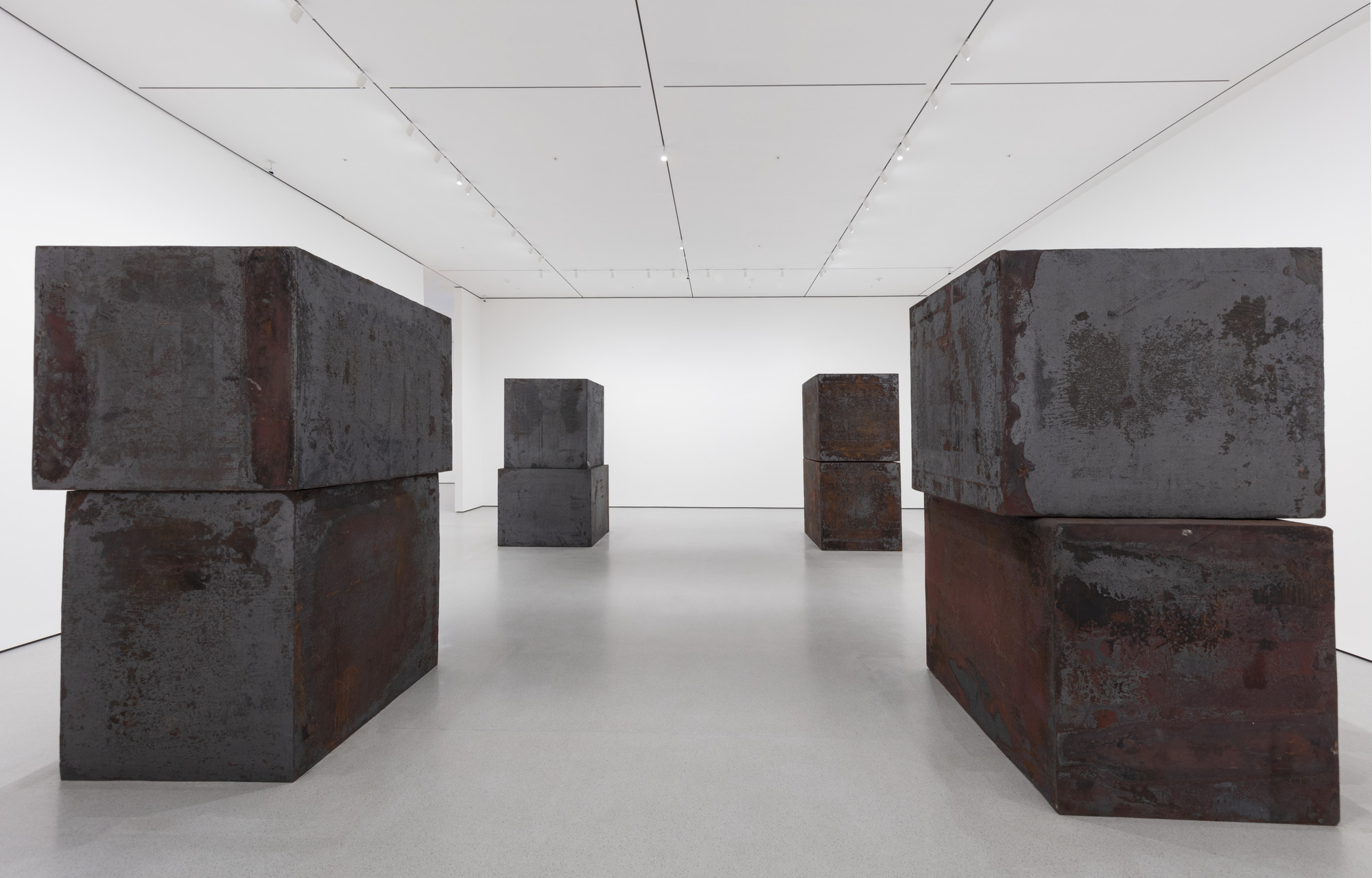 The image shows a gallery with large, dark metal blocks arranged in pairs, one stacked on top of the other. The predominant colors are dark gray and reddish-brown, with a light gray floor and white walls. This is an AI generated description. To give feedback, email ai@moma.org.